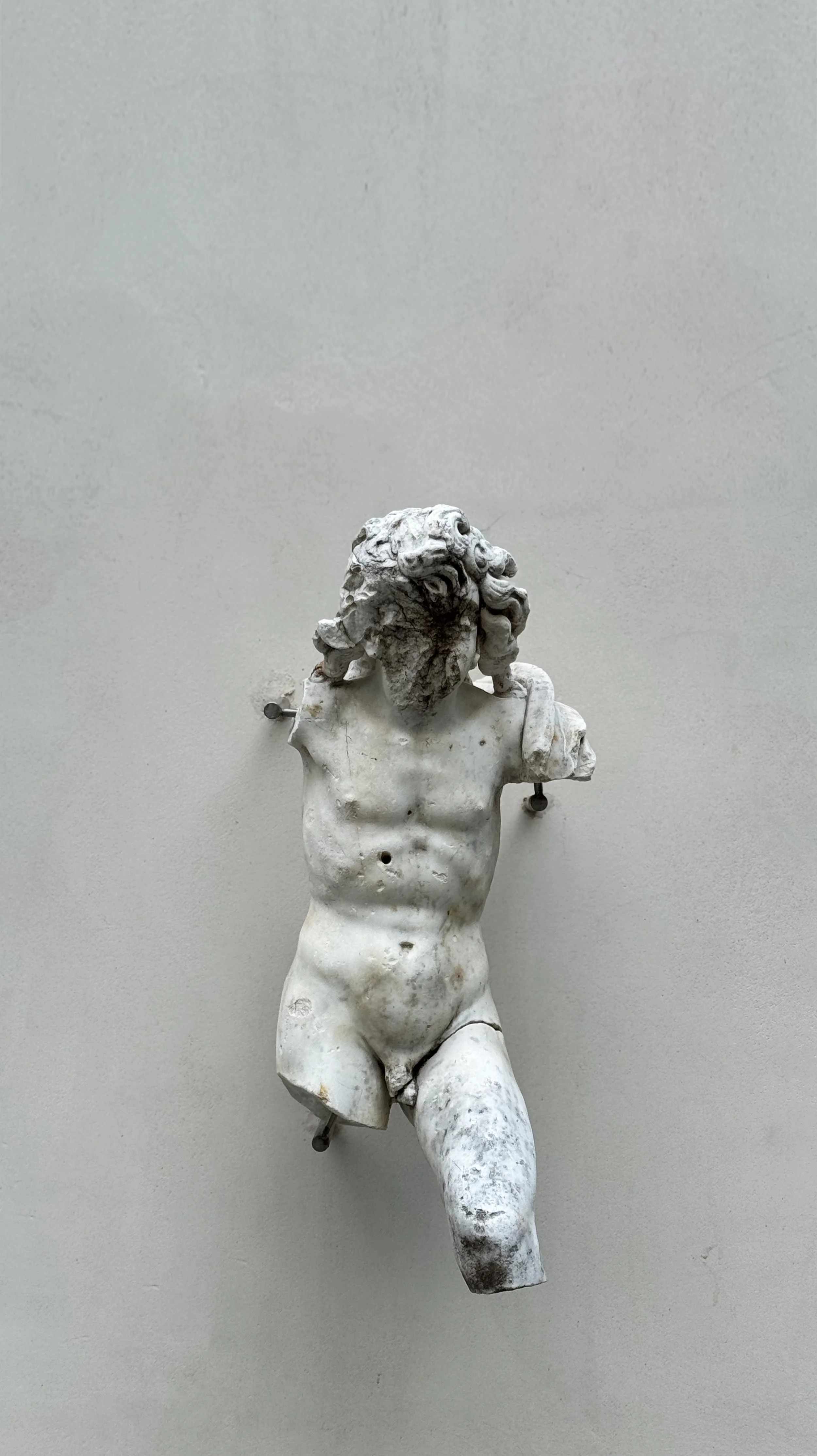 Fragment of ancient marble sculpture depicting the upper torso and head of a male figure, with some parts missing or damaged, mounted on a plain wall.