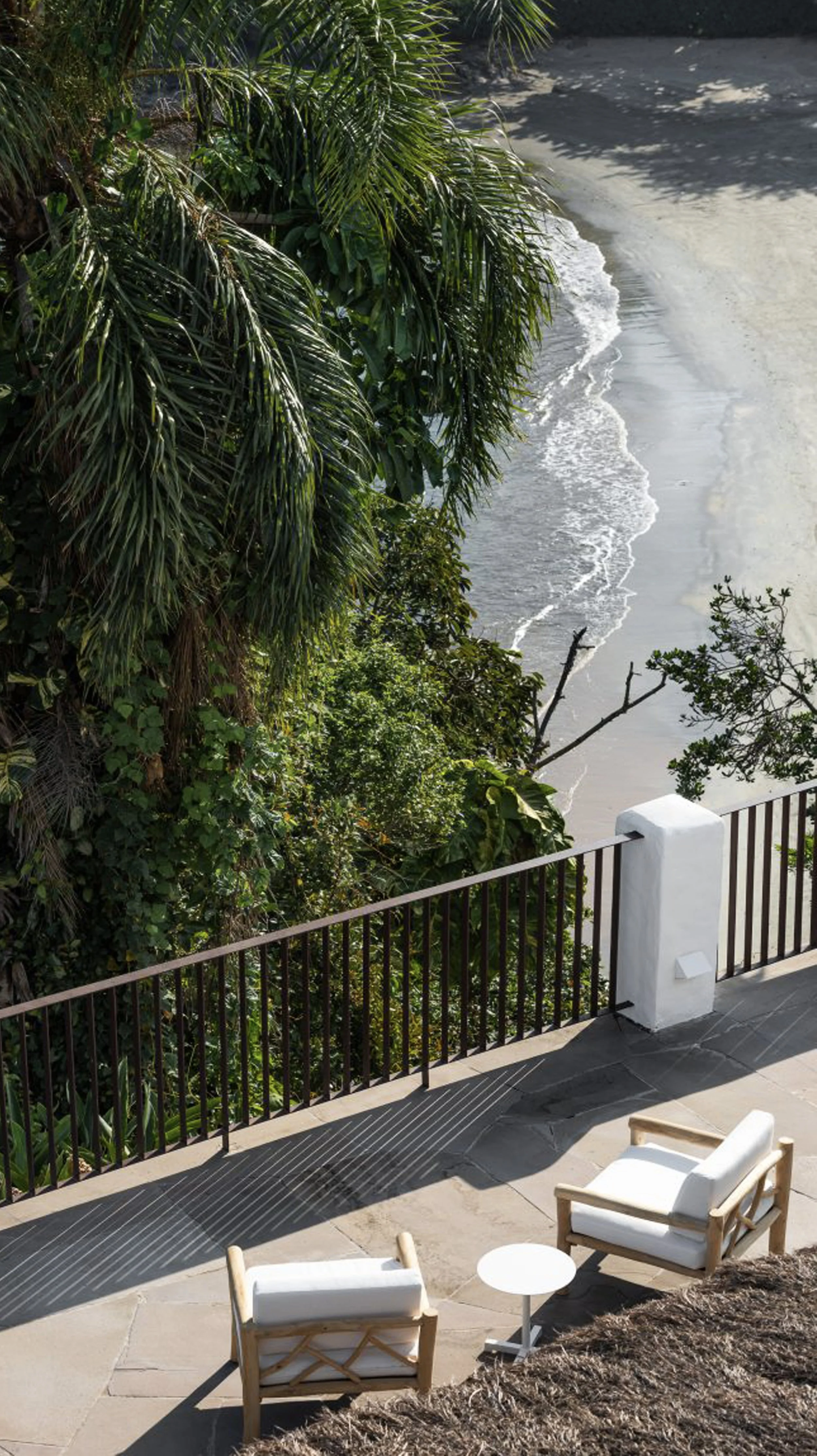 Two outdoor chairs with white cushions and a small round white table on a patio overlooking a beach with sand and ocean waves. Dense green foliage on the left side of the image.