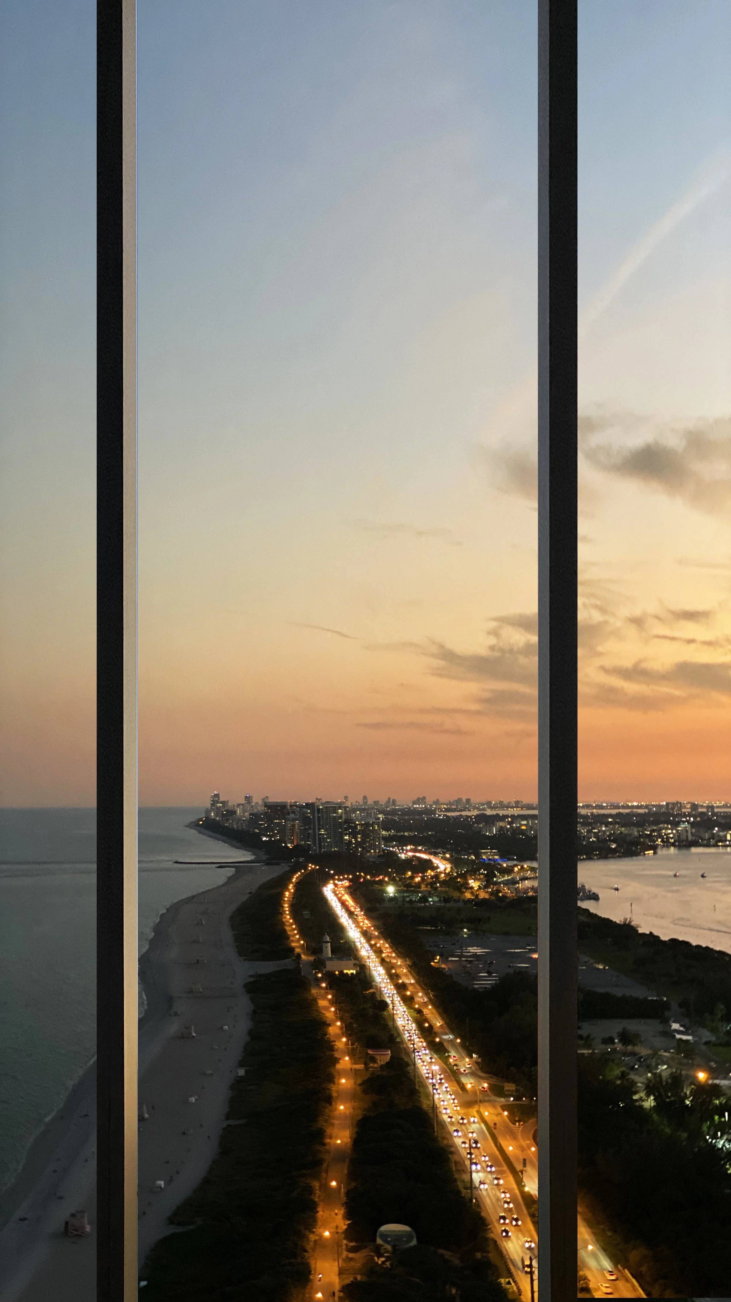 View of a city, beach, and ocean at sunset through window bars, with city lights and traffic on the road.