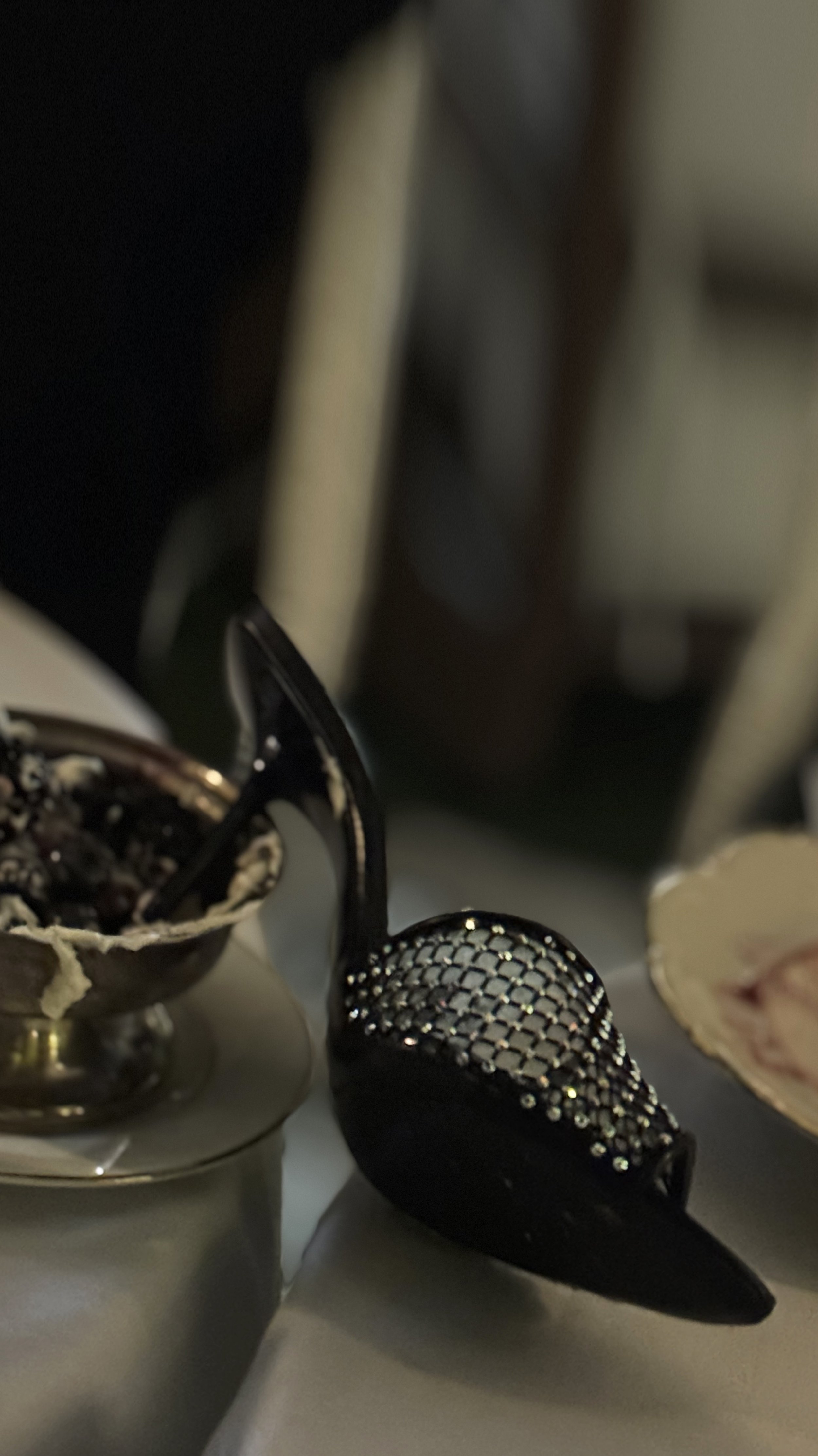 Close-up of a vintage silver spoon resting on a reflective surface, with a decorative black item adorned with rhinestones or crystals nearby, in a dimly lit setting.