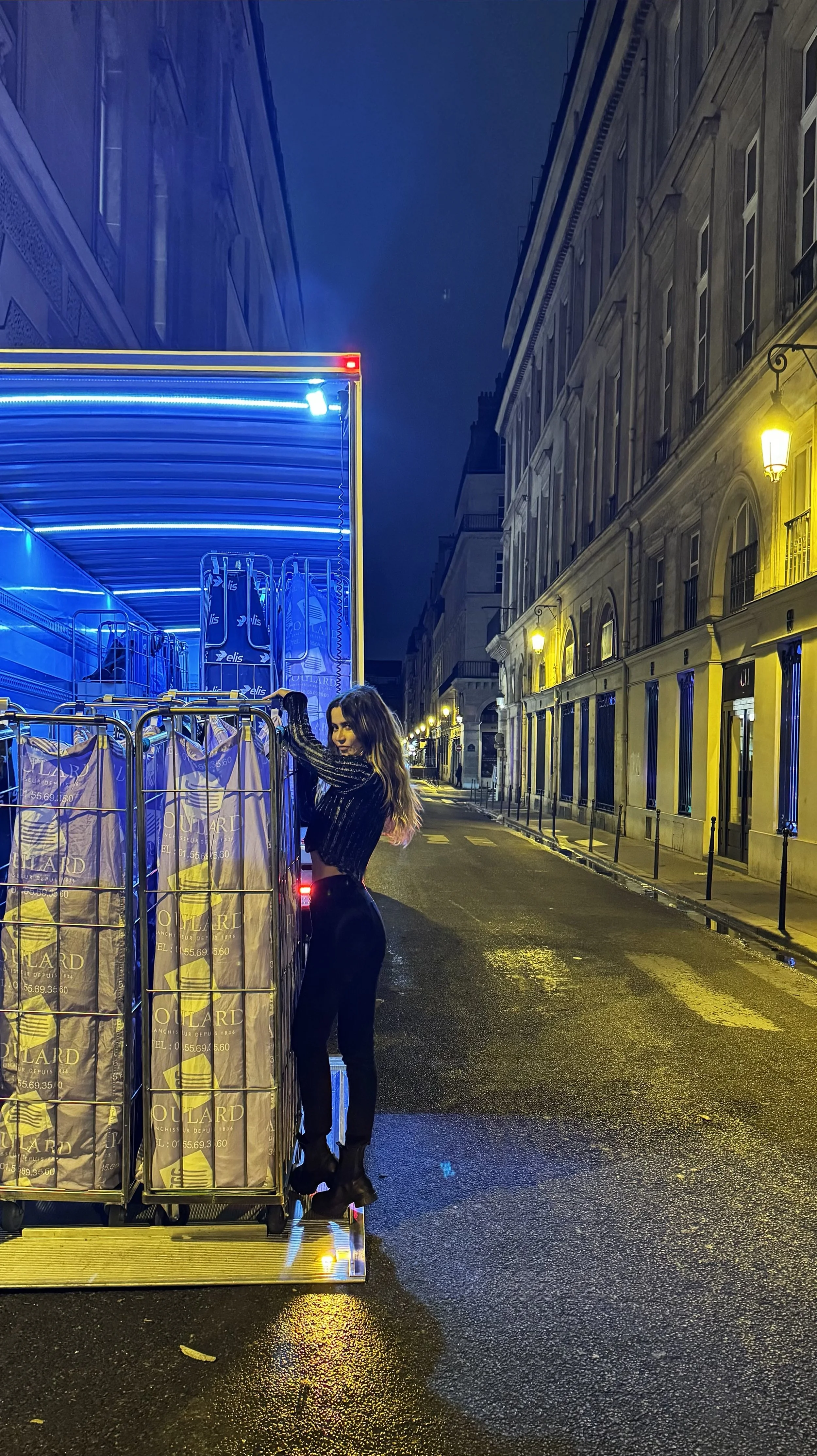 A woman in black outfit loading shopping bags onto a blue delivery truck on a wet city street at night, with old buildings and streetlights illuminated in the background.