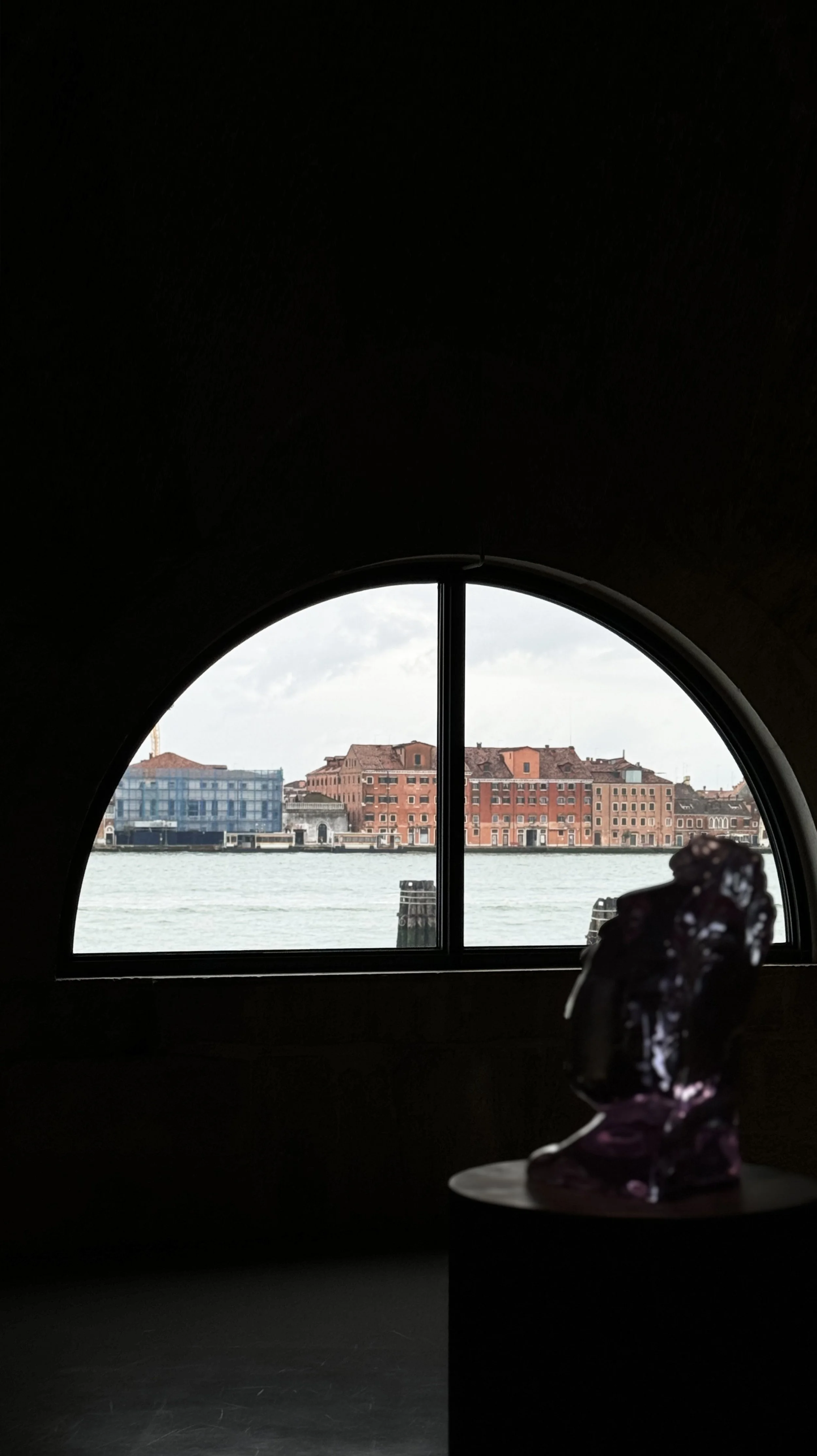 View of a canal with buildings across the water, seen through a rounded window, with a sculpture of a child's head silhouette in the foreground.