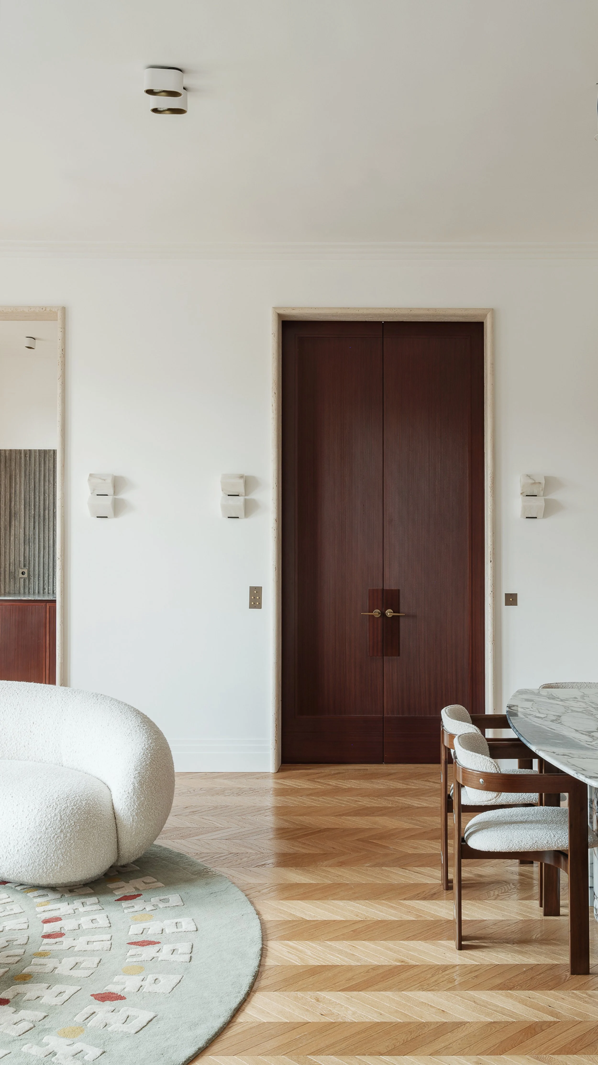 Interior view of a modern living space with wooden floors, a round beige seating, and a marble dining table with chairs. Centered is a large wooden double door on a white wall, with minimalist light fixtures on either side.
