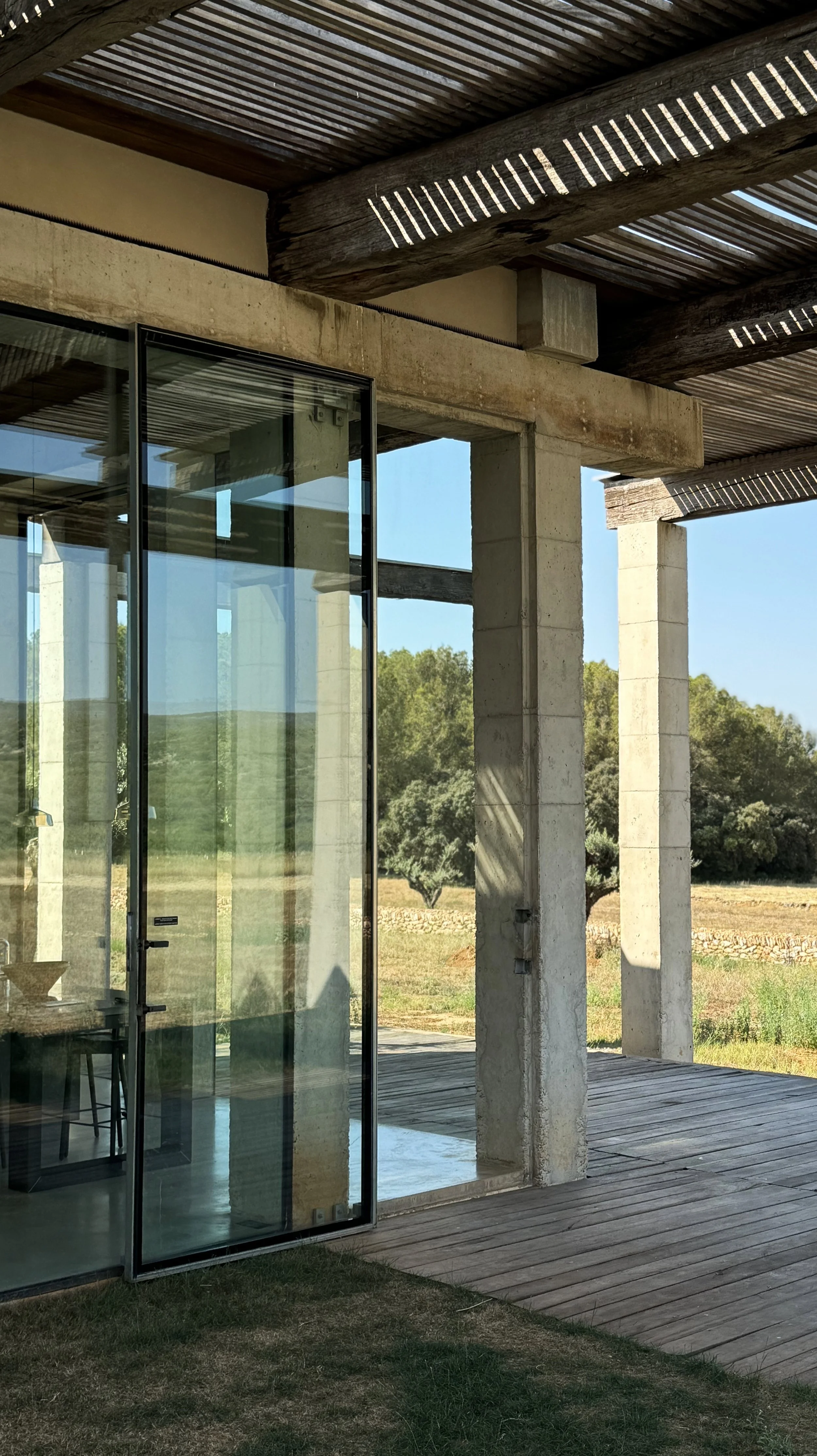 Exterior view of a modern house with large glass sliding doors, concrete pillars, and wooden ceiling beams, surrounded by a grassy lawn and trees in the background.