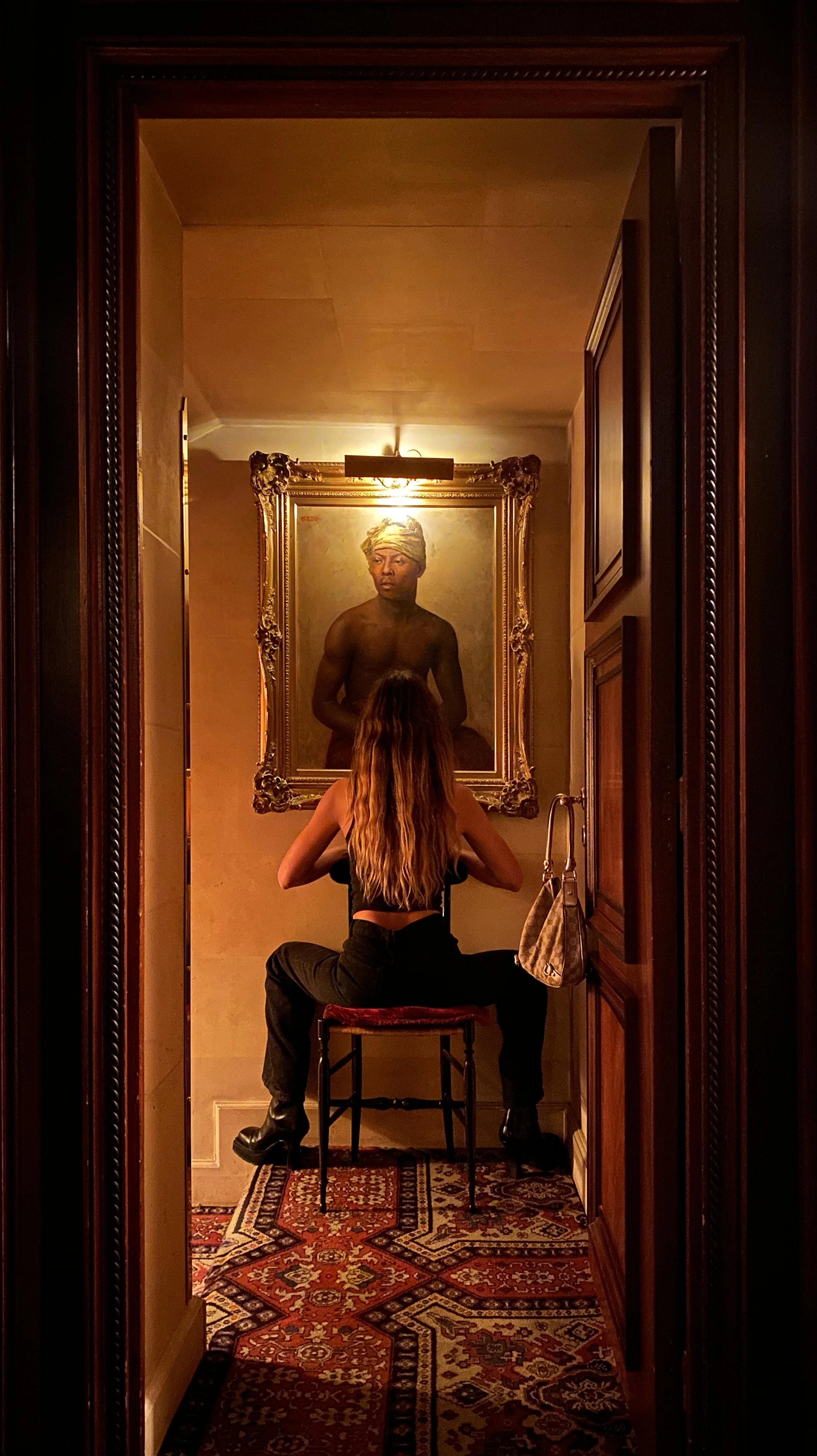 A woman with long wavy hair sitting on a chair in a small, warmly lit room, facing a large portrait of a shirtless man with a towel wrapped around his head, enclosed in an ornate frame.