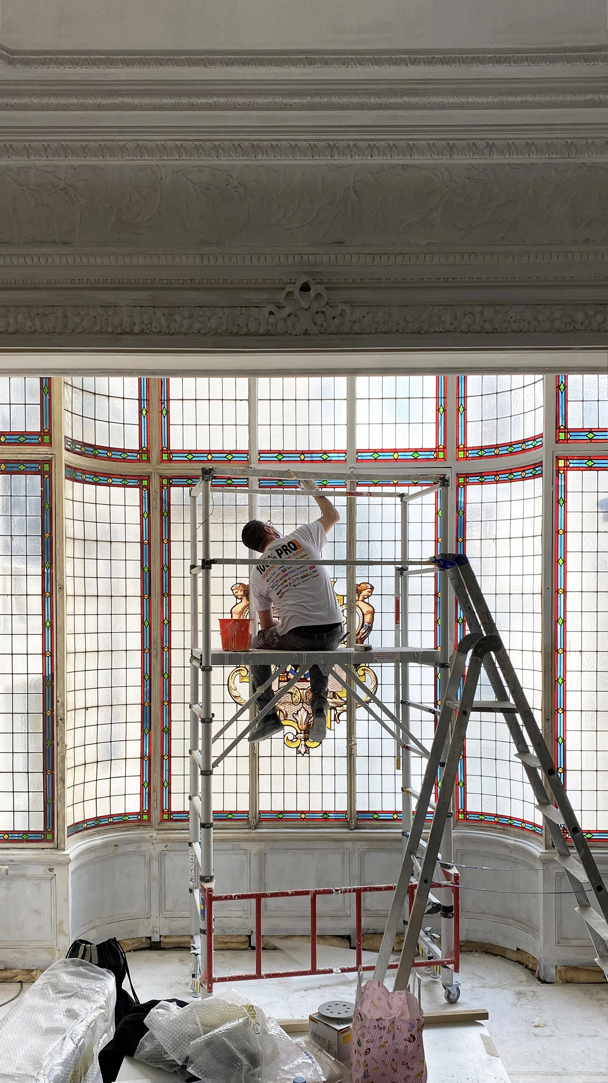A worker on scaffolding restoring a large stained glass window with red, green, and blue decorative elements in a historic building.
