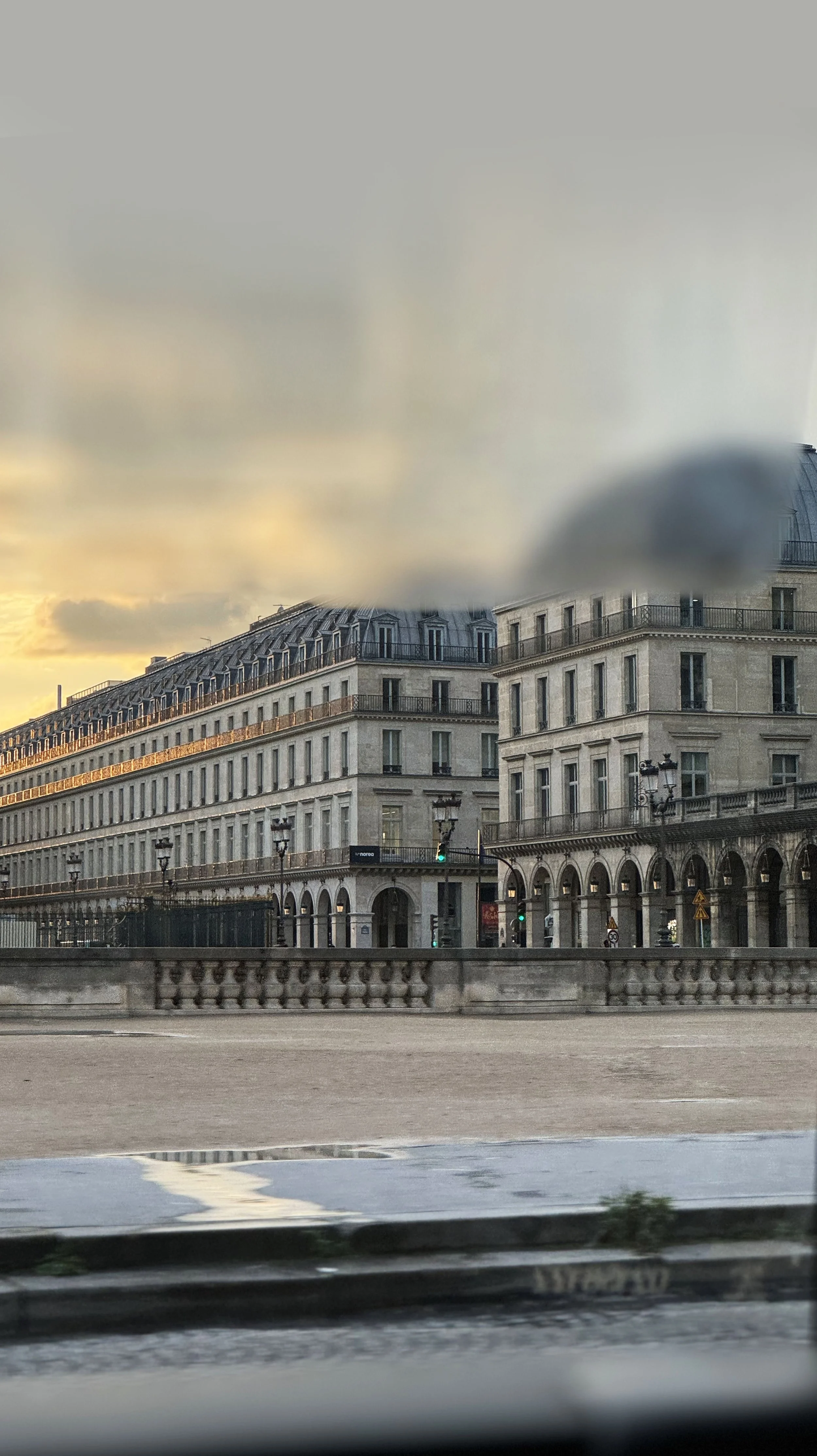 Parisian street view at sunset with classic French architecture, featuring multiple-story buildings with balconies, arched windows, and a dome-shaped roof in the background. A cloudy sky with soft sunlight is visible.