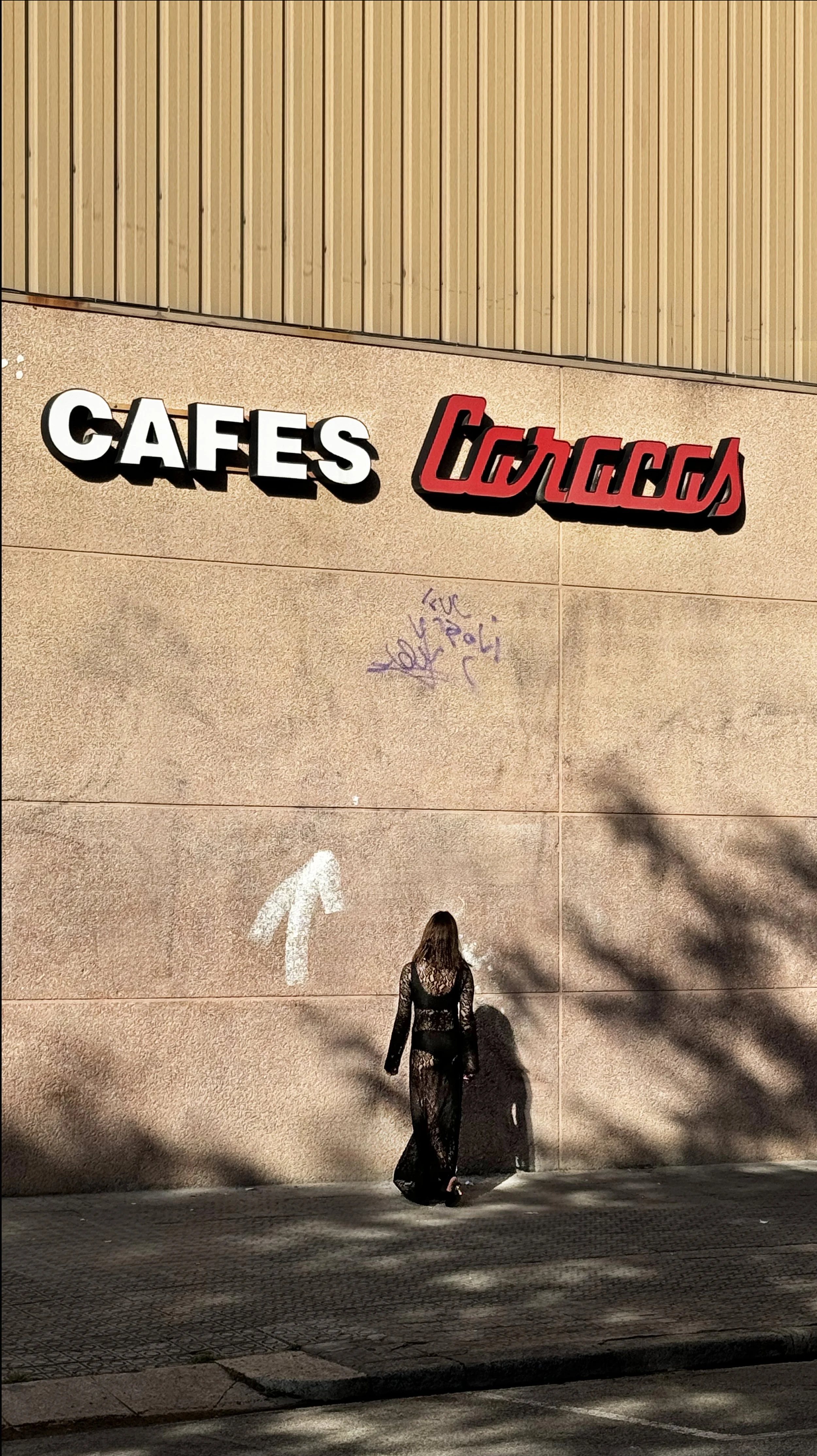 A woman in a black lace dress walking along a sidewalk near a beige wall with graffiti, shadows of tree branches, and a sign that reads Political Cafes.