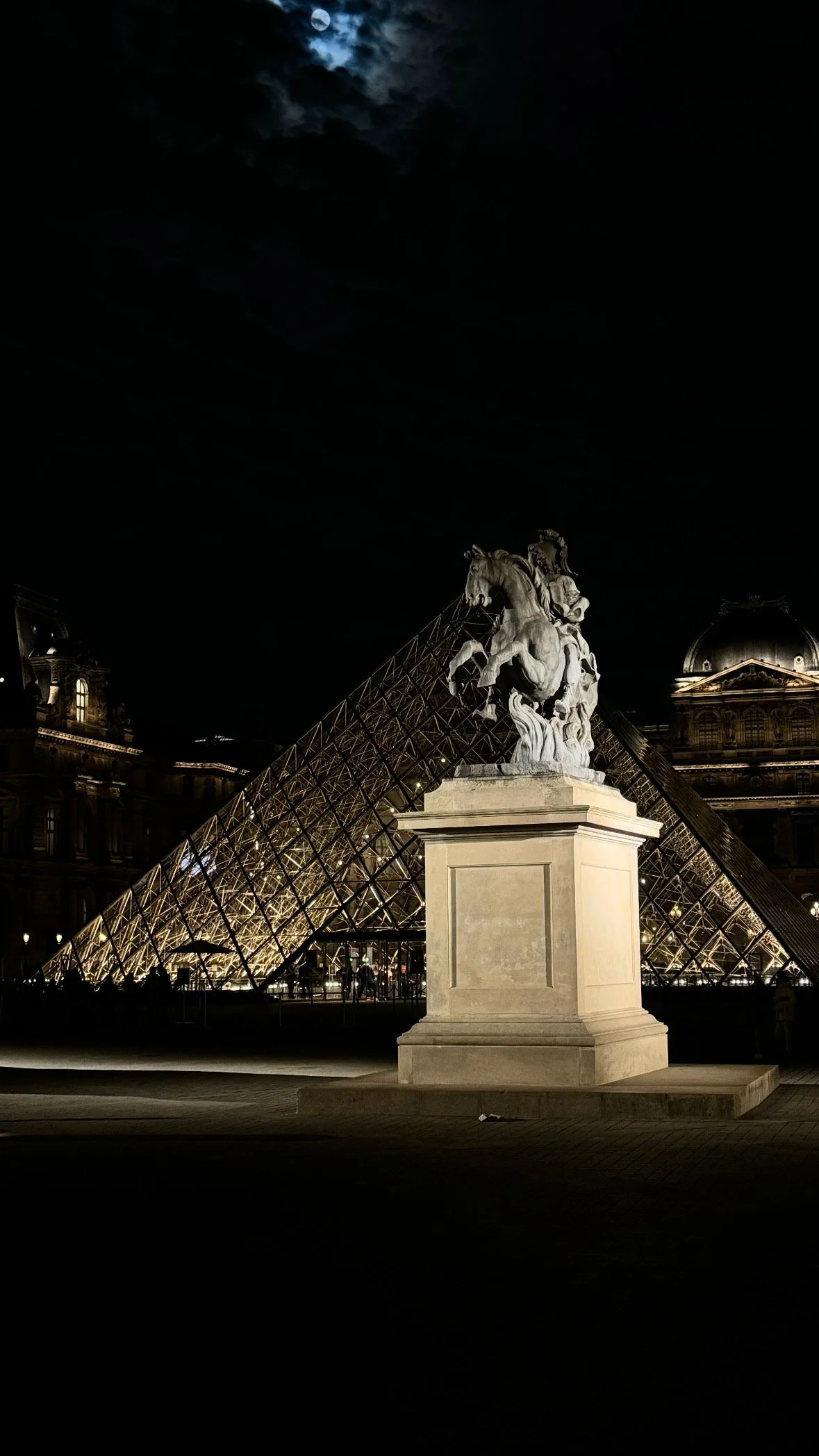 Nighttime view of the Louvre Museum in Paris with the illuminated glass pyramid and a stone statue of a horse and rider in the foreground, under a cloudy moonlit sky.