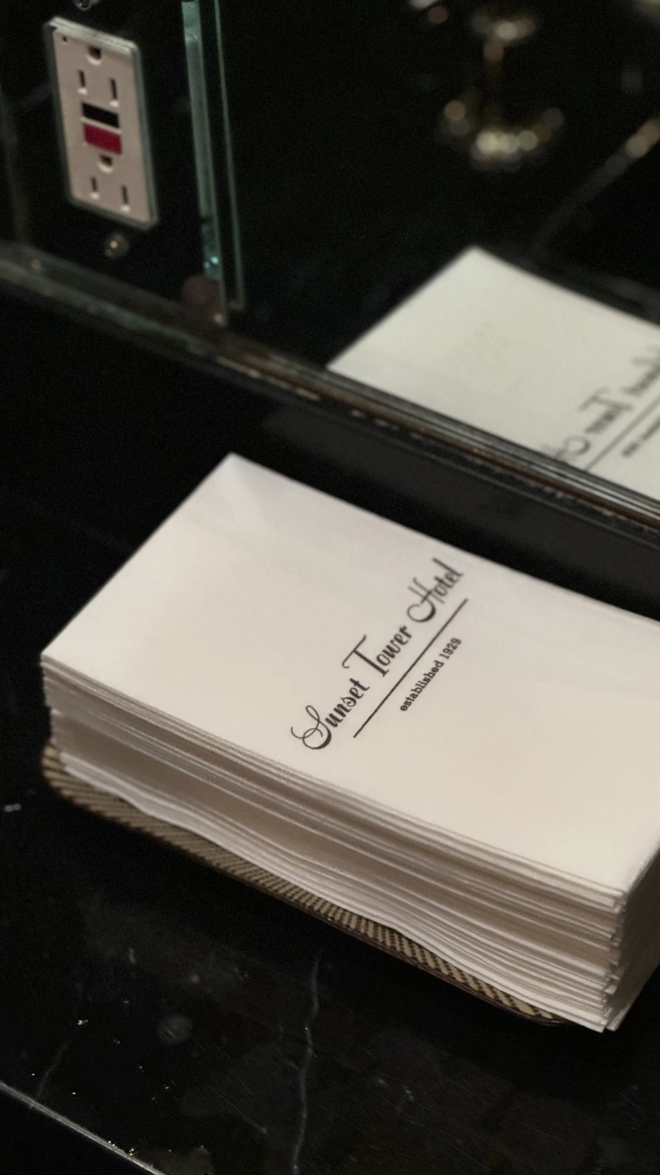 Stack of napkins with Sunset Tower Hotel printed on top, placed near a mirror and an electrical outlet.