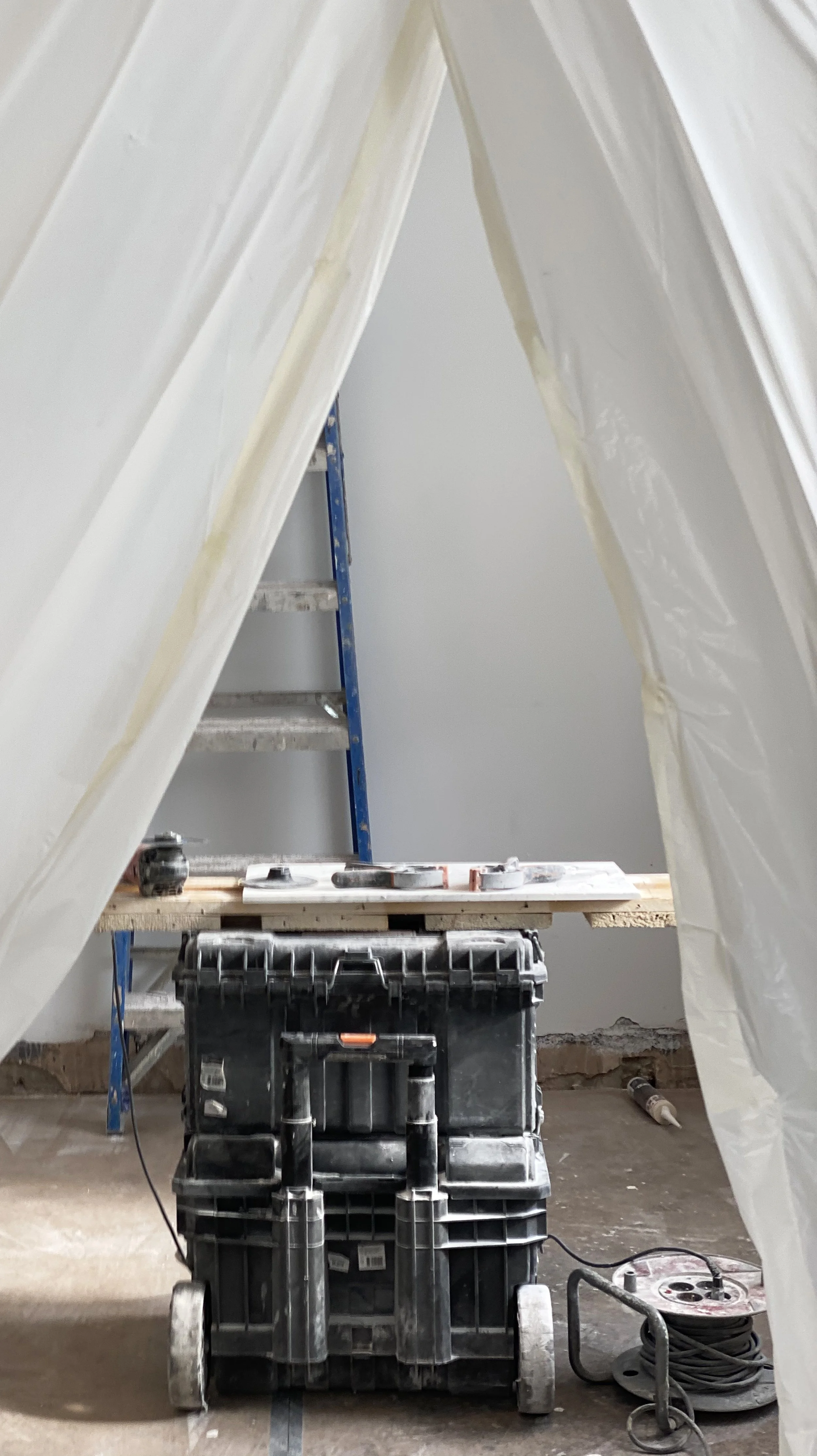 Workshop with a stack of black tool boxes, a wooden work table with tools on it, a blue ladder, and a plastic sheet forming a tent-like structure.