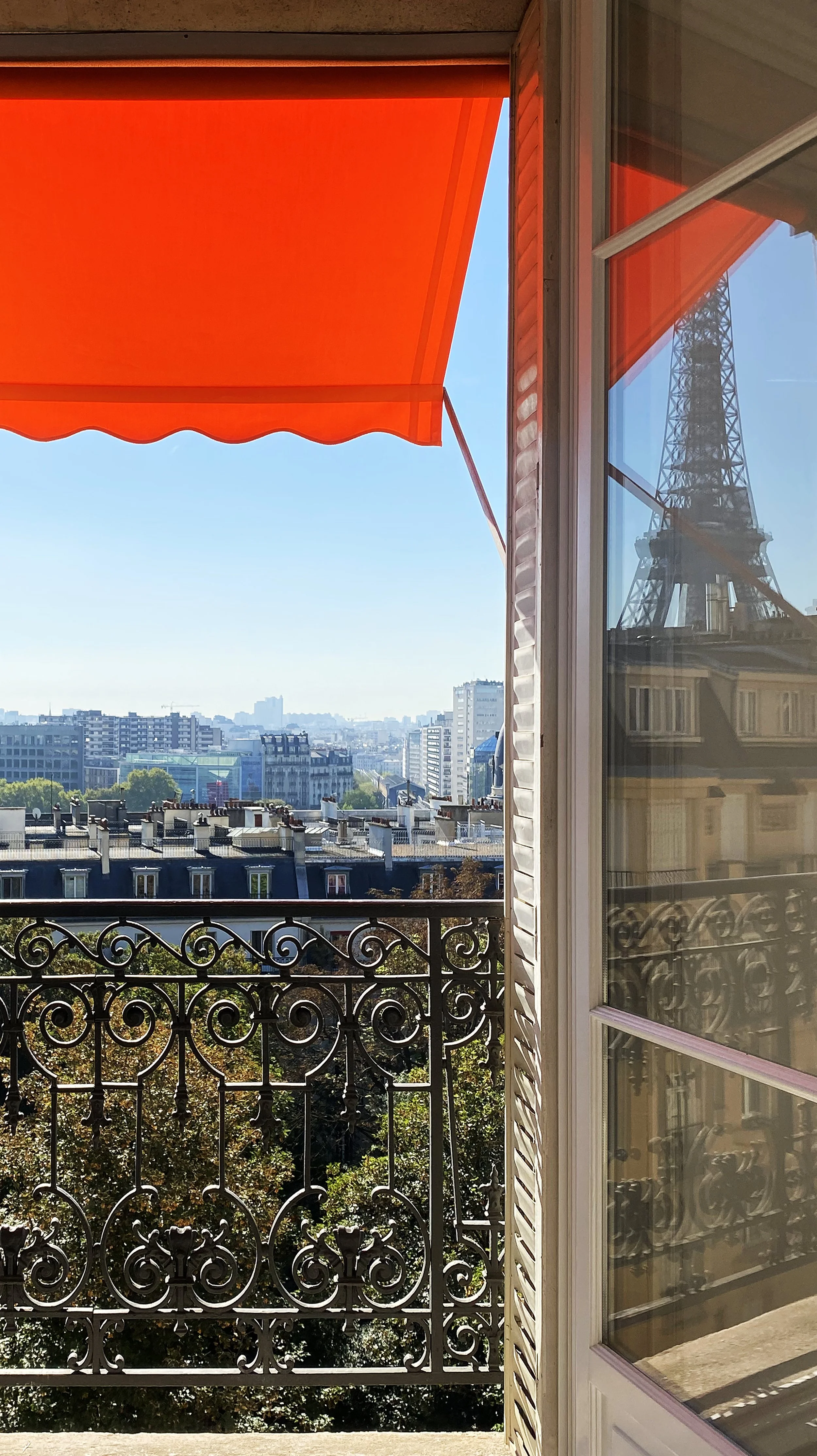 A view from a balcony in Paris showing the Eiffel Tower reflected in the window and a vibrant red awning.