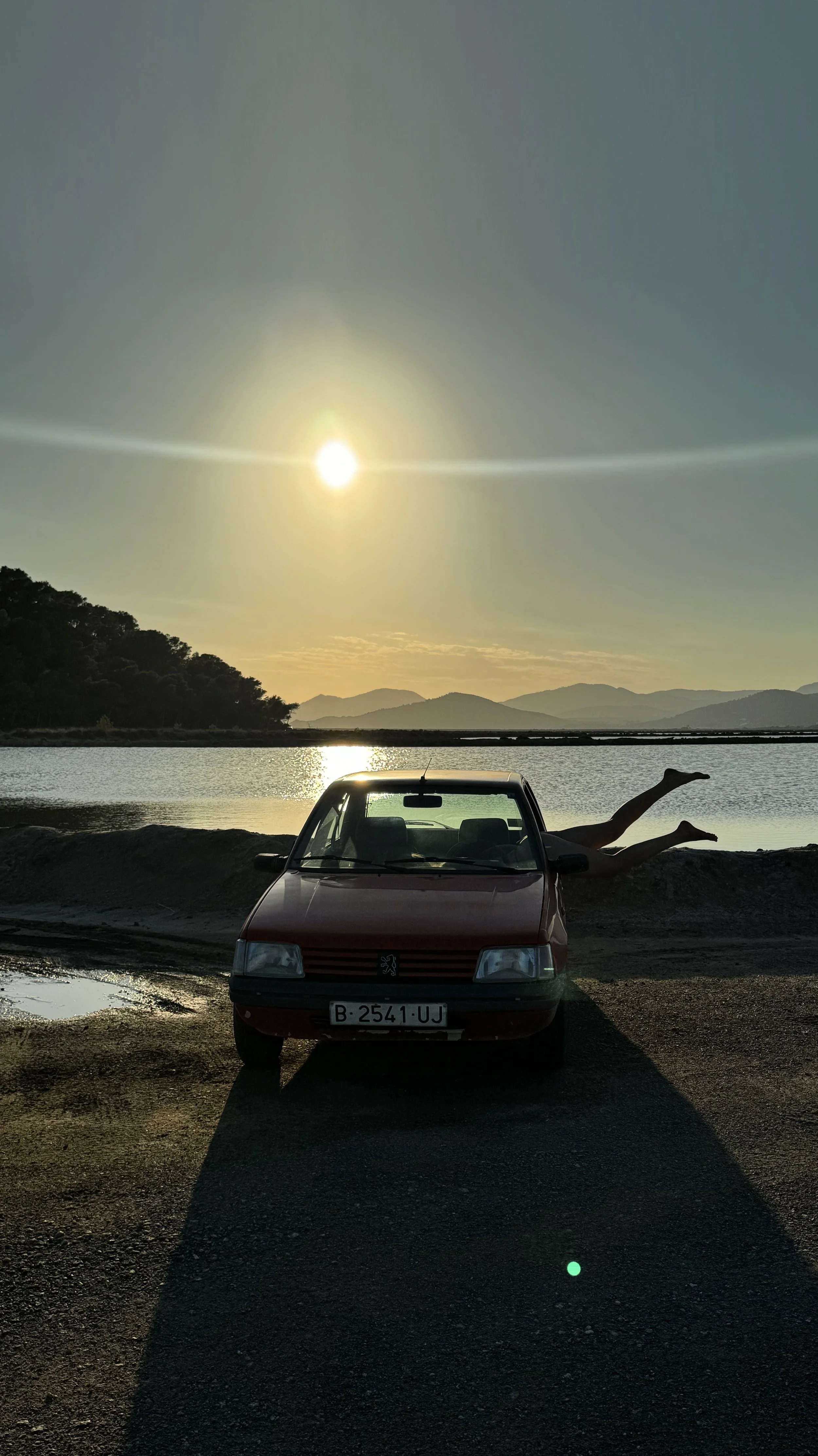 A red vintage car parked near a body of water during sunset, with a person lying on the ground next to it, legs playfully stretched out behind the car, and mountains in the background.