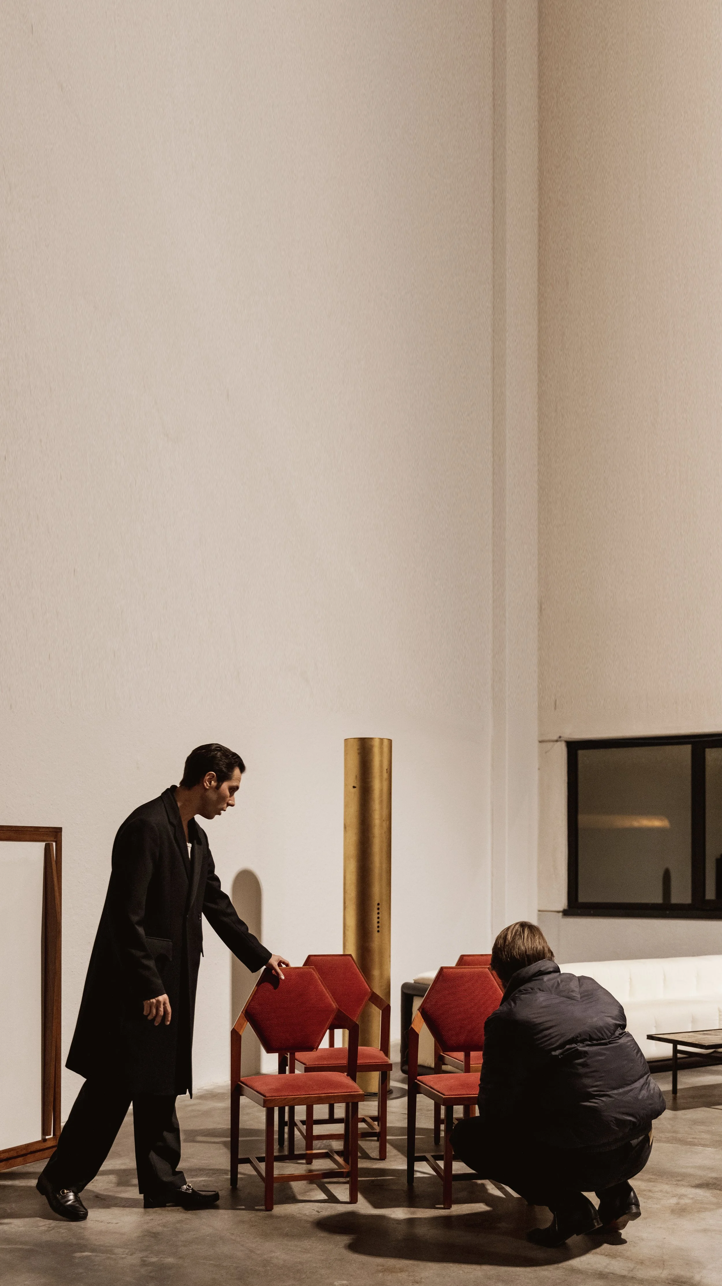 Two men are discussing or inspecting red chairs in a modern, minimalistic interior with white walls, a window, and a large golden sculpture or pipe.
