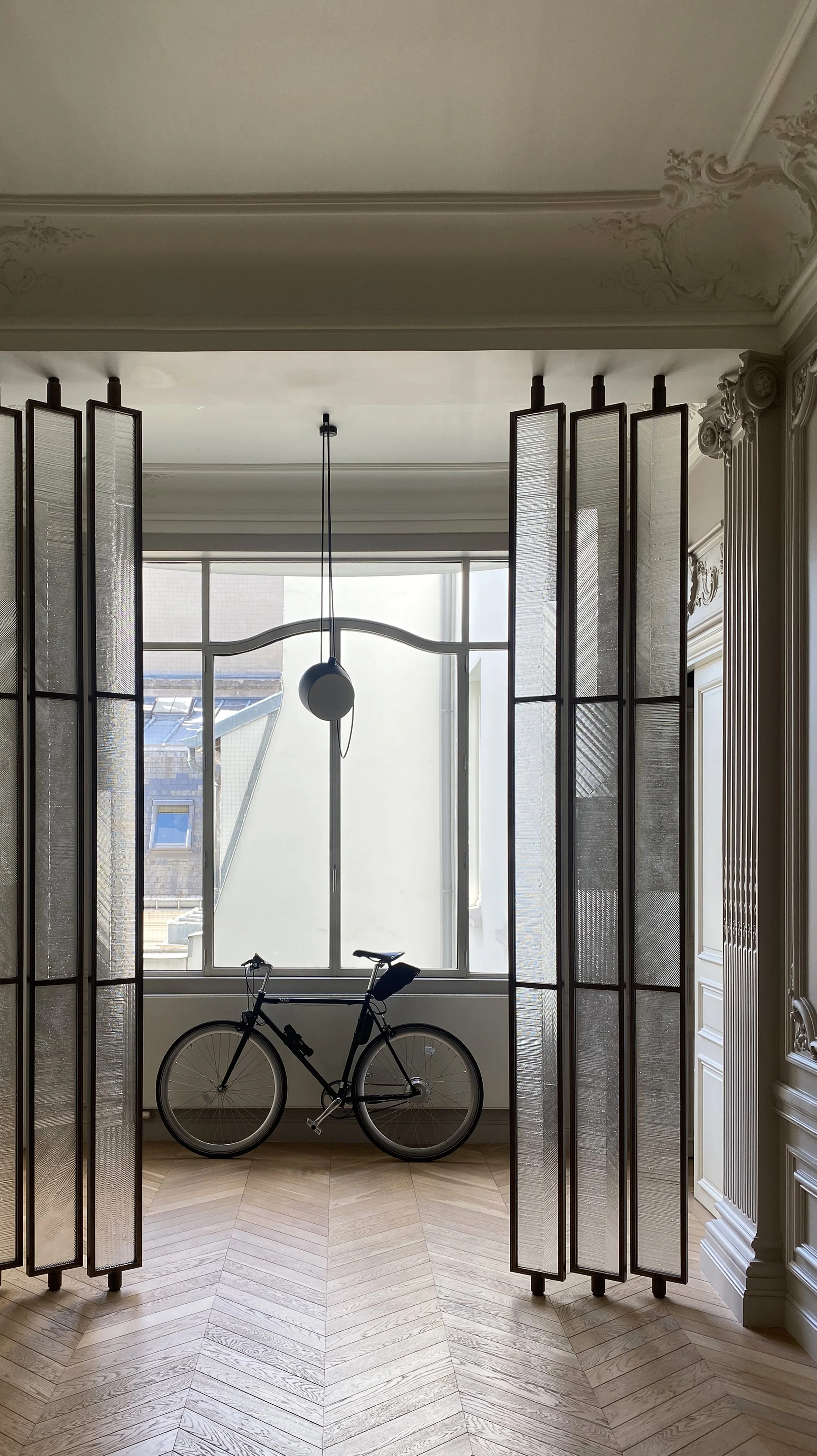 Interior of a room with a vintage style, featuring a large bay window, decorative moldings on the ceiling and walls, a black bicycle leaning against the window, and a modern hanging light fixture.