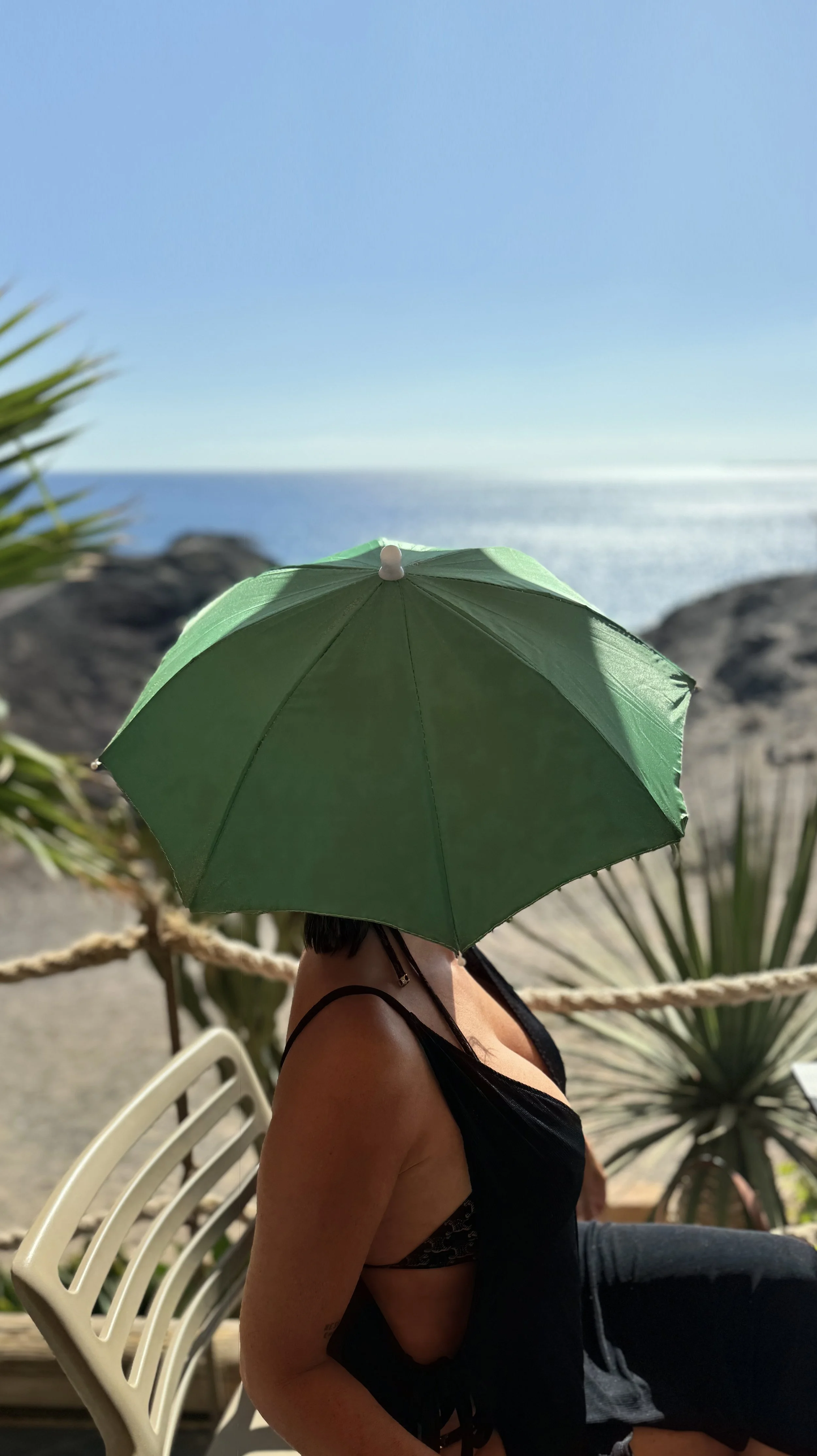 Woman sitting outdoors with an ocean view, holding a green umbrella, wearing a black top, and surrounded by plants.