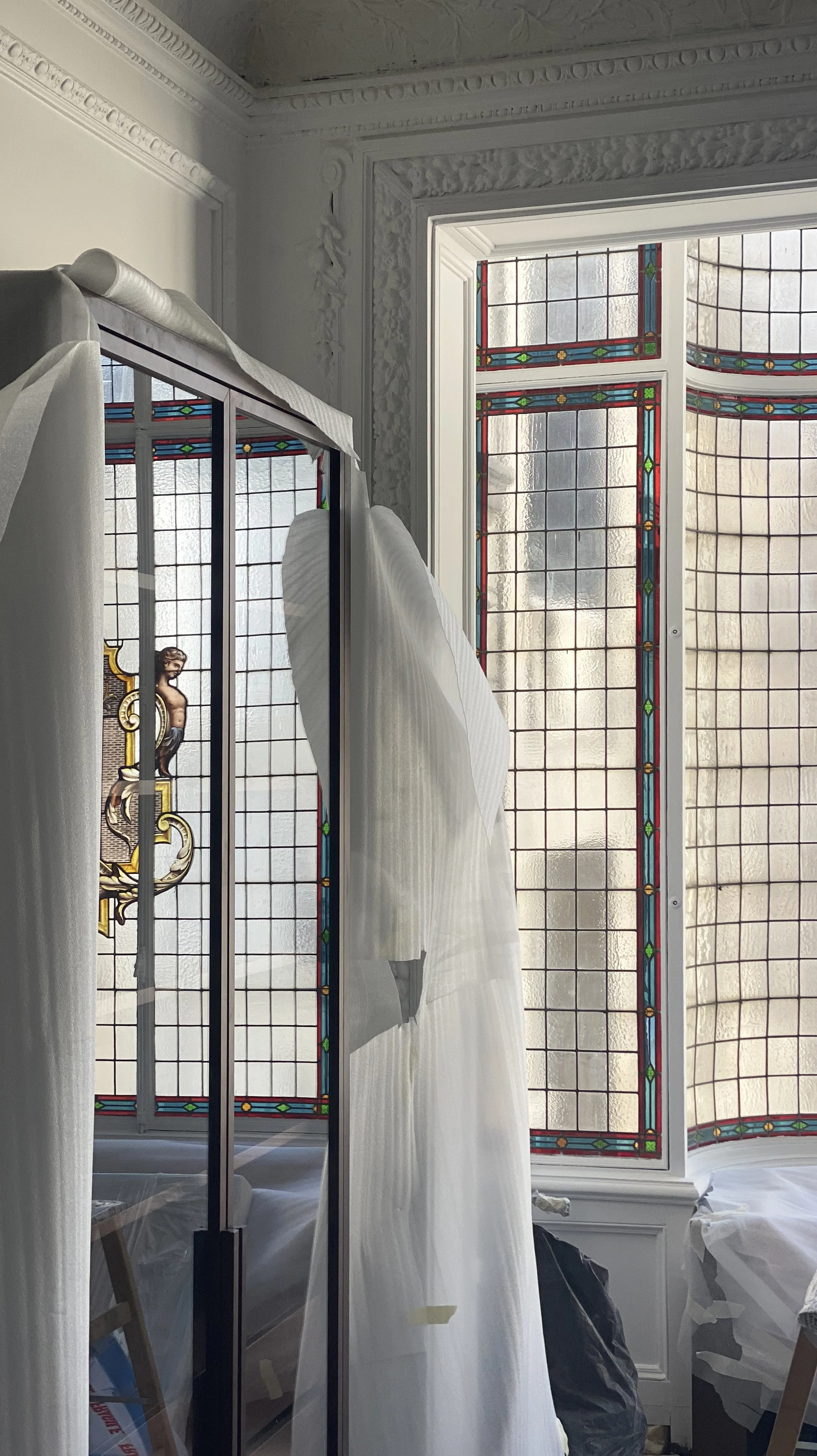 An interior room with large stained glass windows and a mirror reflecting part of the room. There is a white curtain or fabric hanging and some furniture and objects partially visible.