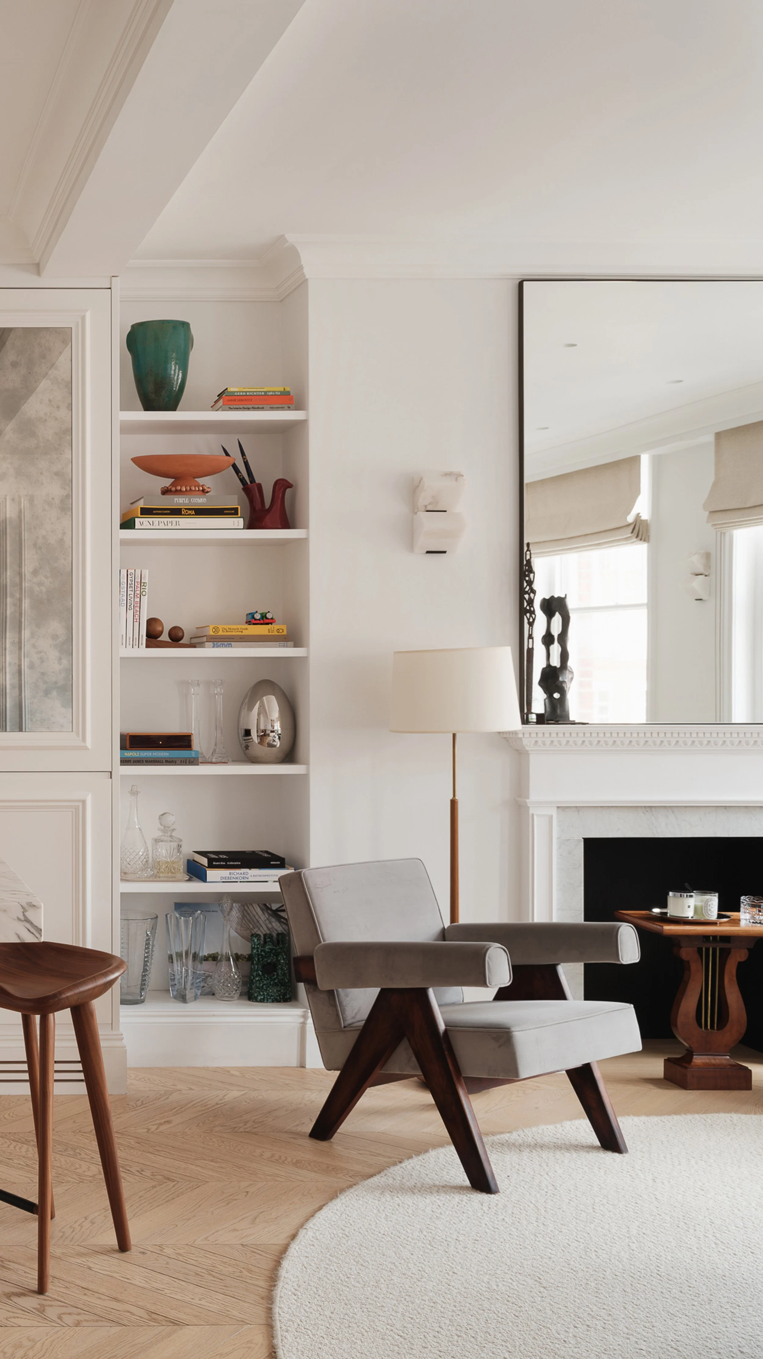 Modern living room with white walls, a chandelier, a gray armchair, a white bookshelf with decorative items, a floor lamp, a fireplace, and a large mirror.