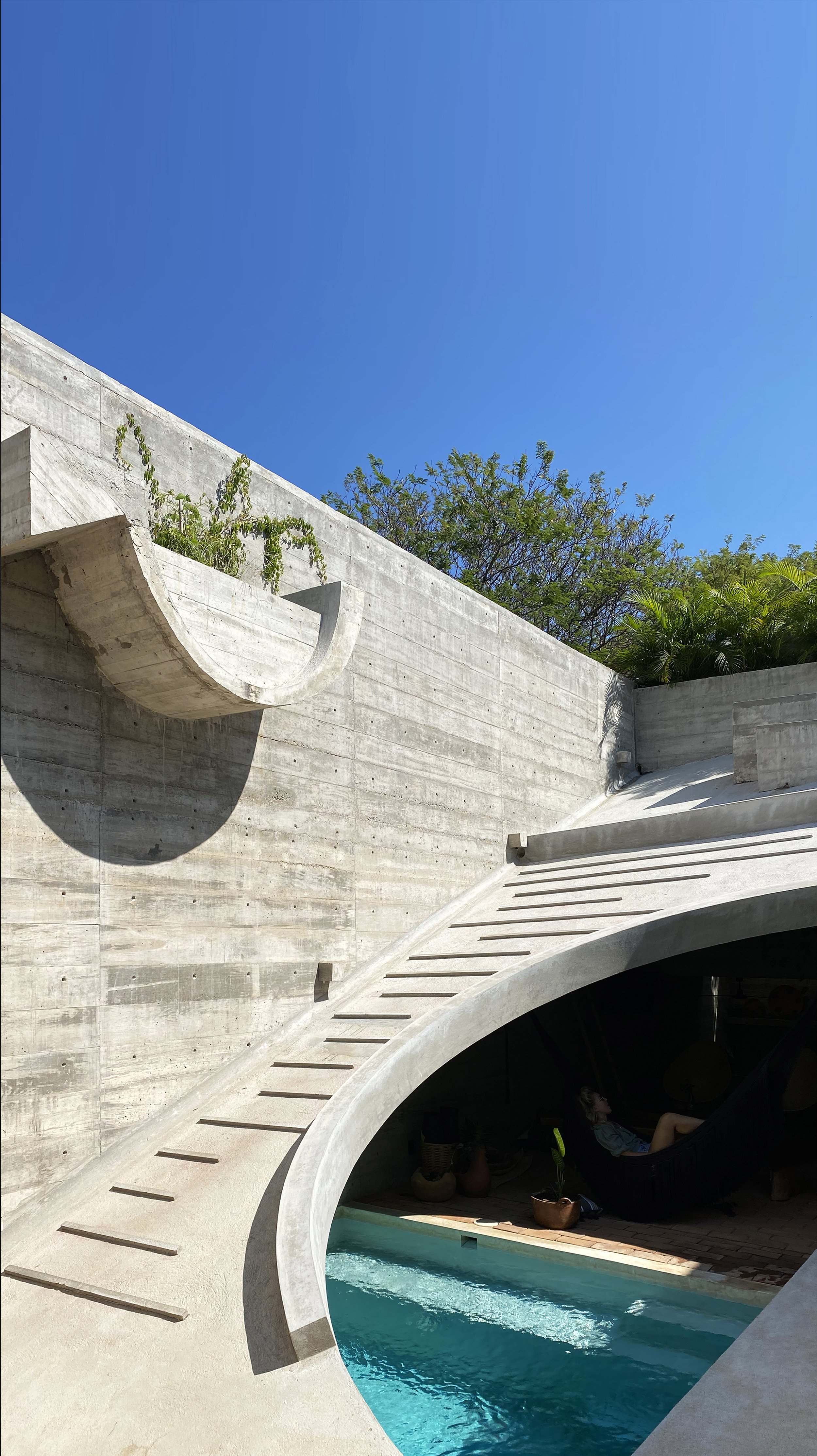 Modern architectural concrete structure with an outdoor pool, curved staircase, and a person resting in a black hammock under an arch, set against a bright blue sky and green trees.