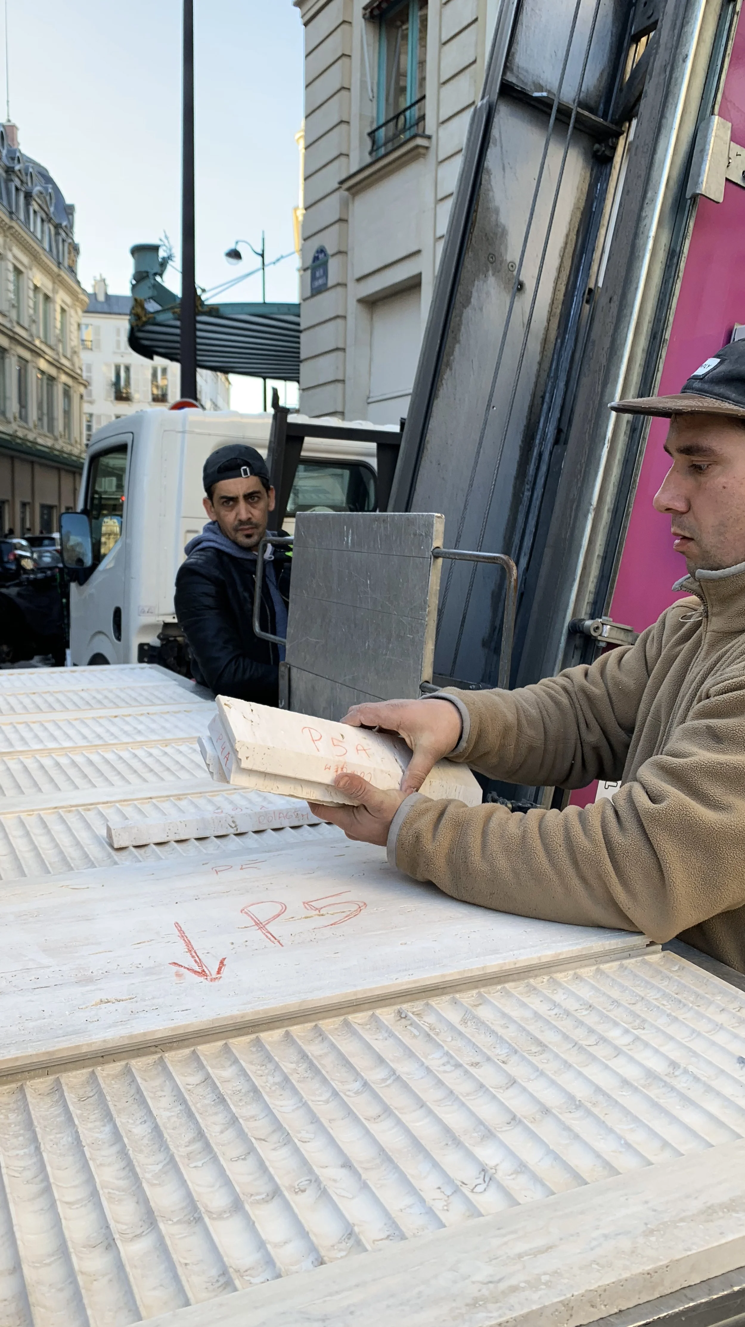 Two men working outdoors, one is handling white tiles or slabs, the other is looking on. They are on a city street, with buildings and parked cars visible in the background.