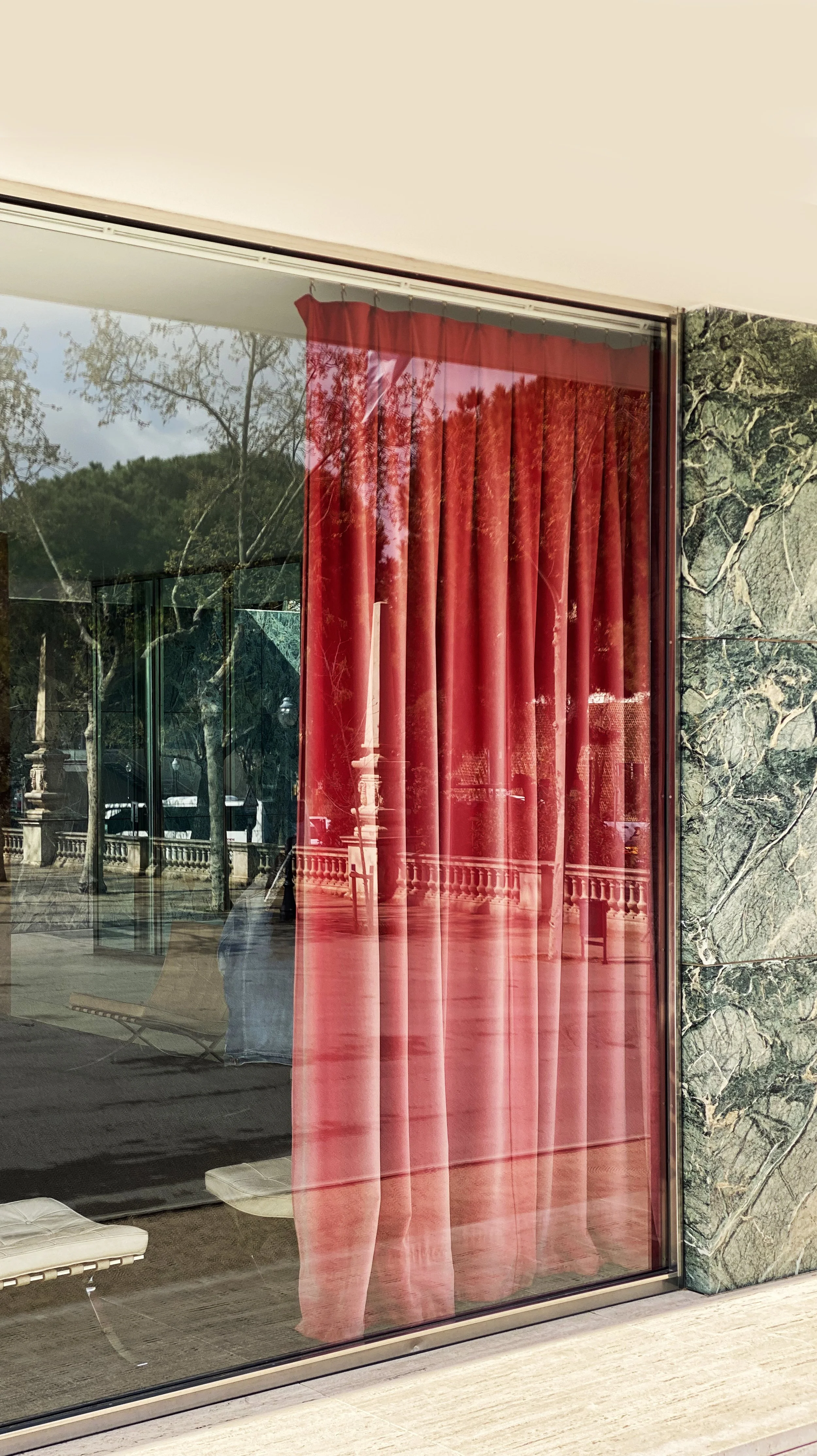 Reflection of trees and a person taking a photo in a large glass window with red curtains inside.