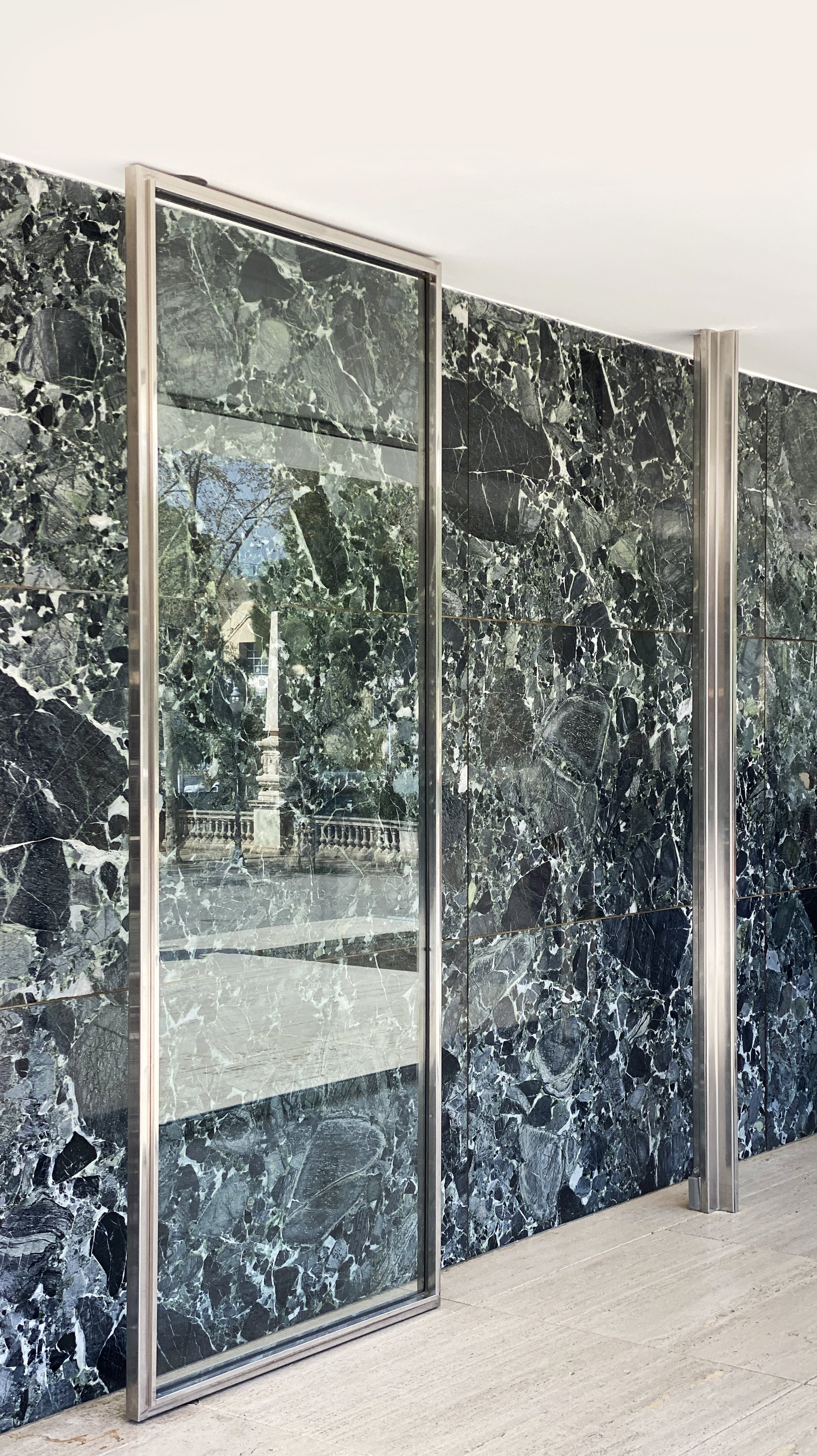 Dark green marble wall with a large glass panel door and metallic framing, reflecting an outdoor scene with trees and a balustrade.