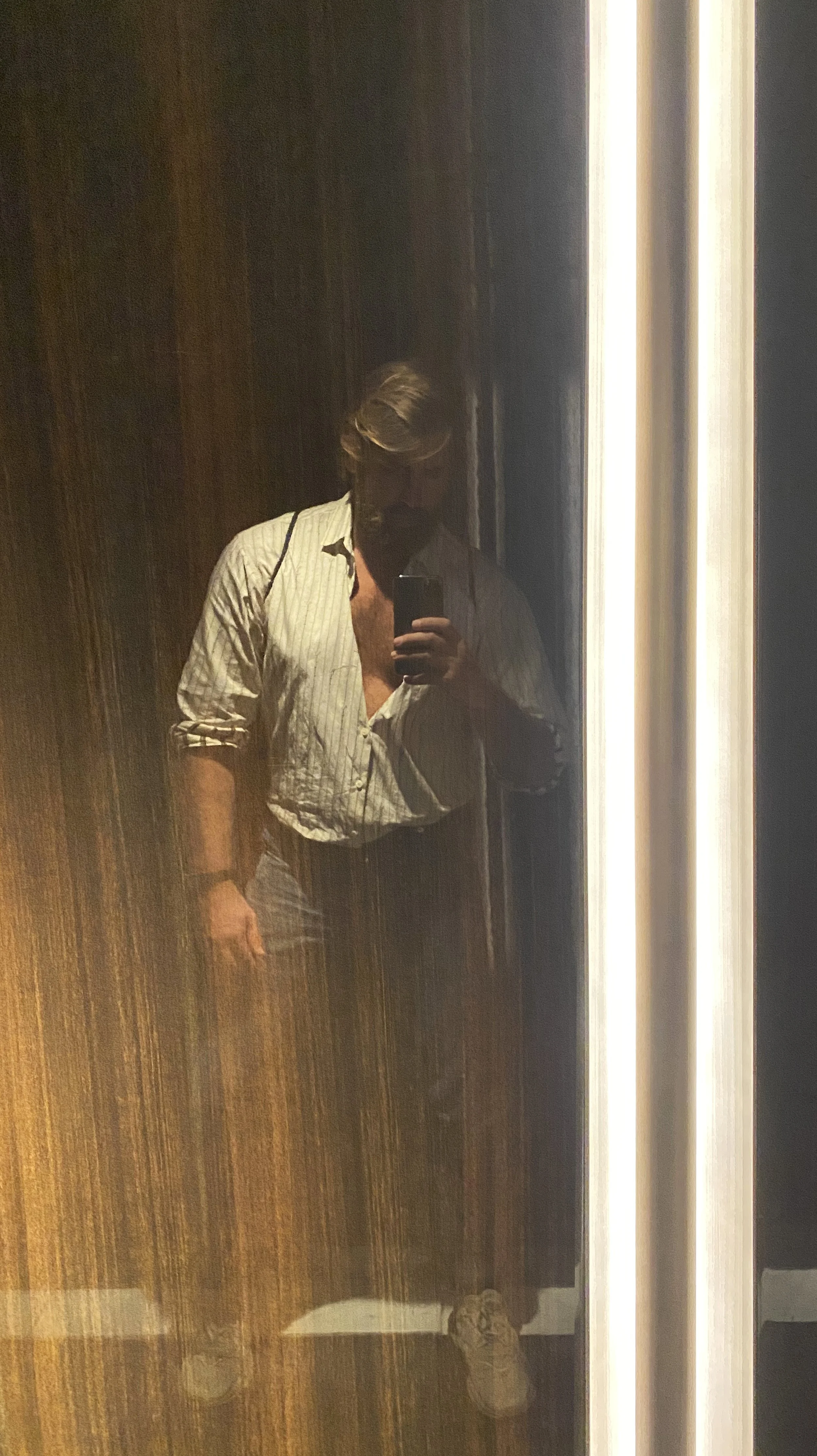 Person taking a mirror selfie in a dimly lit elevator, wearing a striped button-up shirt and gray pants.