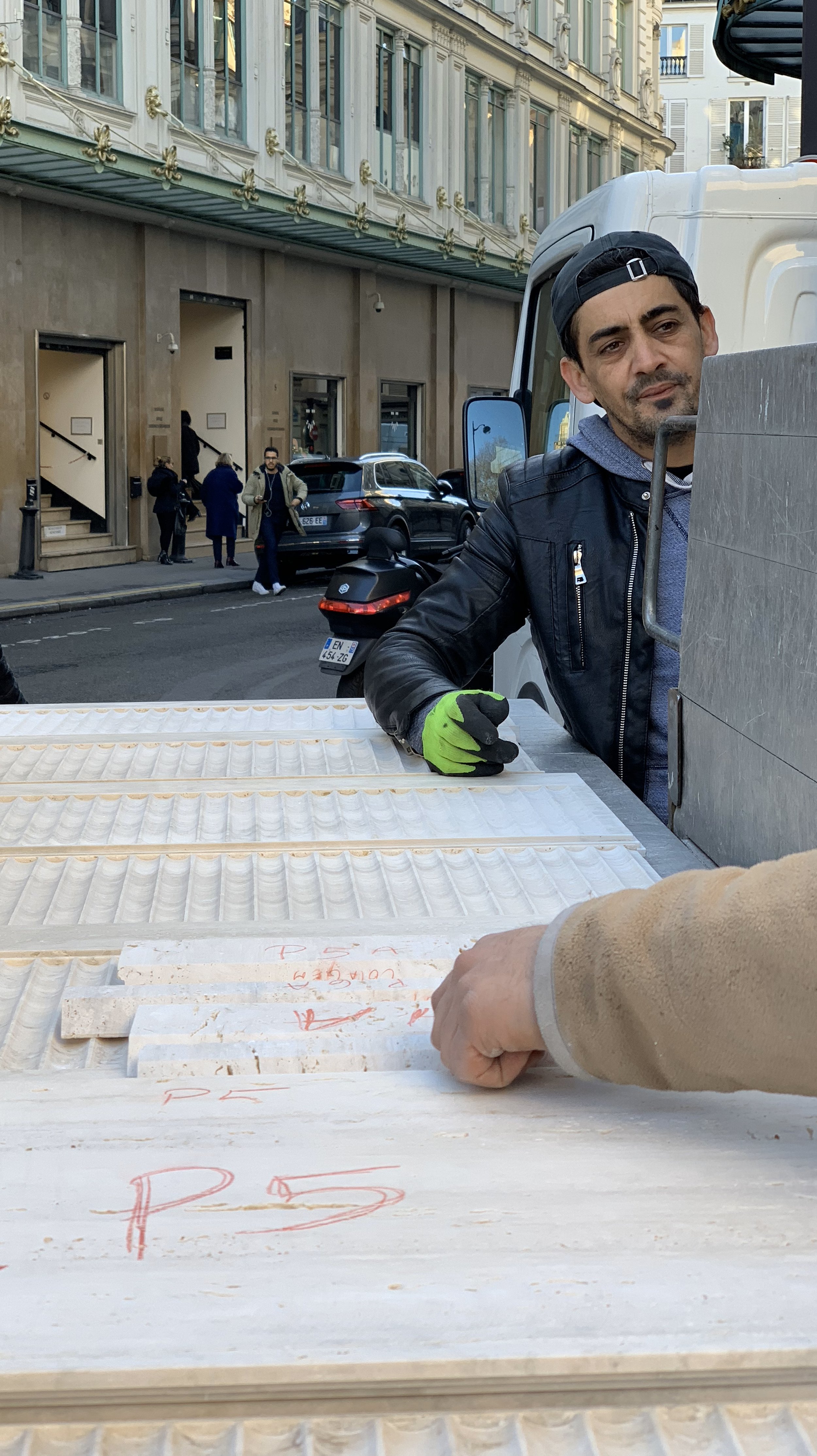 A man with a black cap and black leather jacket stands outdoors on a city street, loading or unloading wooden trays from a truck. Several people are walking on the sidewalk in the background, and parked cars and motorcycles are visible.