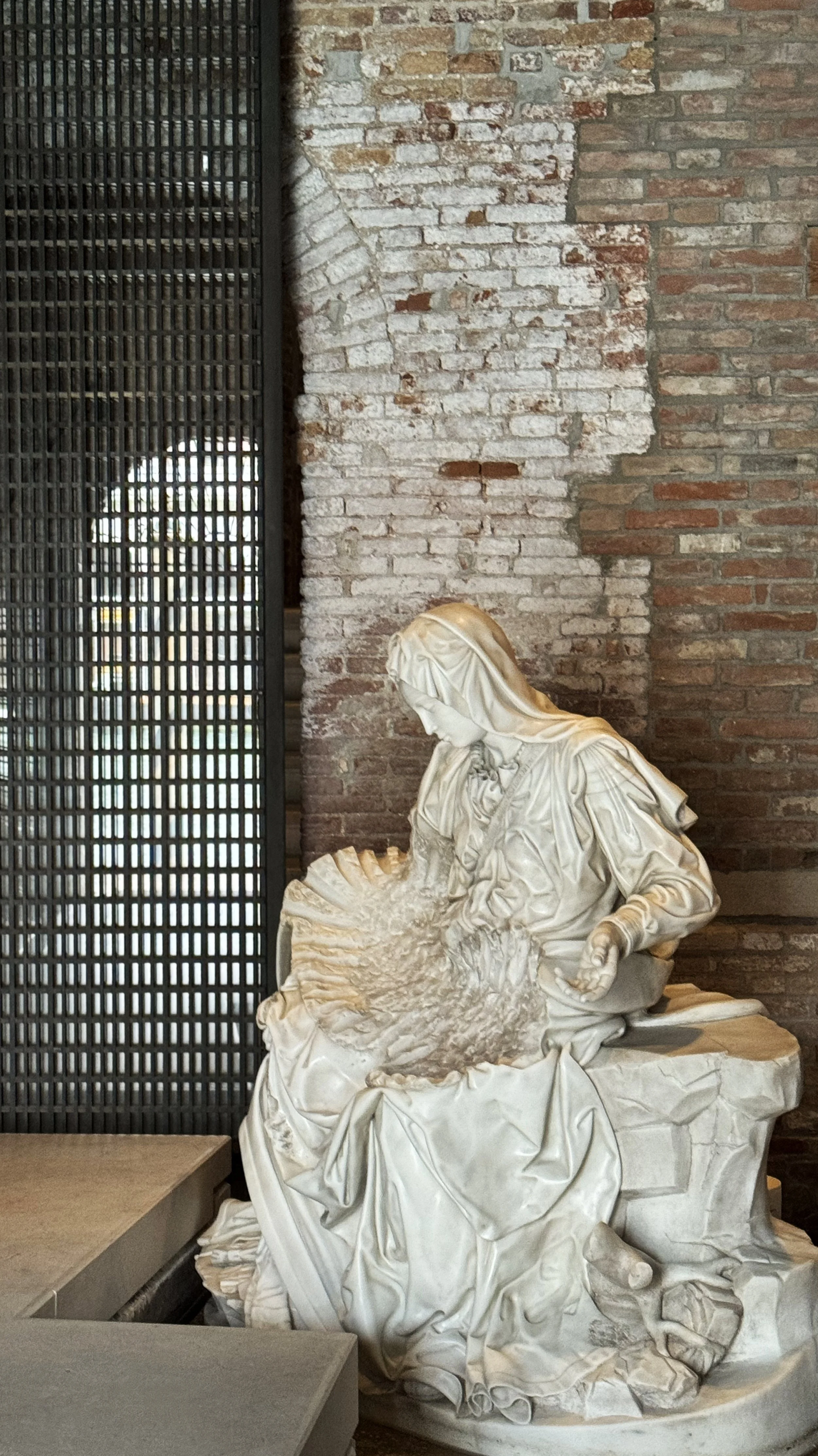 Marble sculpture of a woman with a headscarf, holding a soapy rag, sitting on a cloth-covered surface, against a brick wall background.
