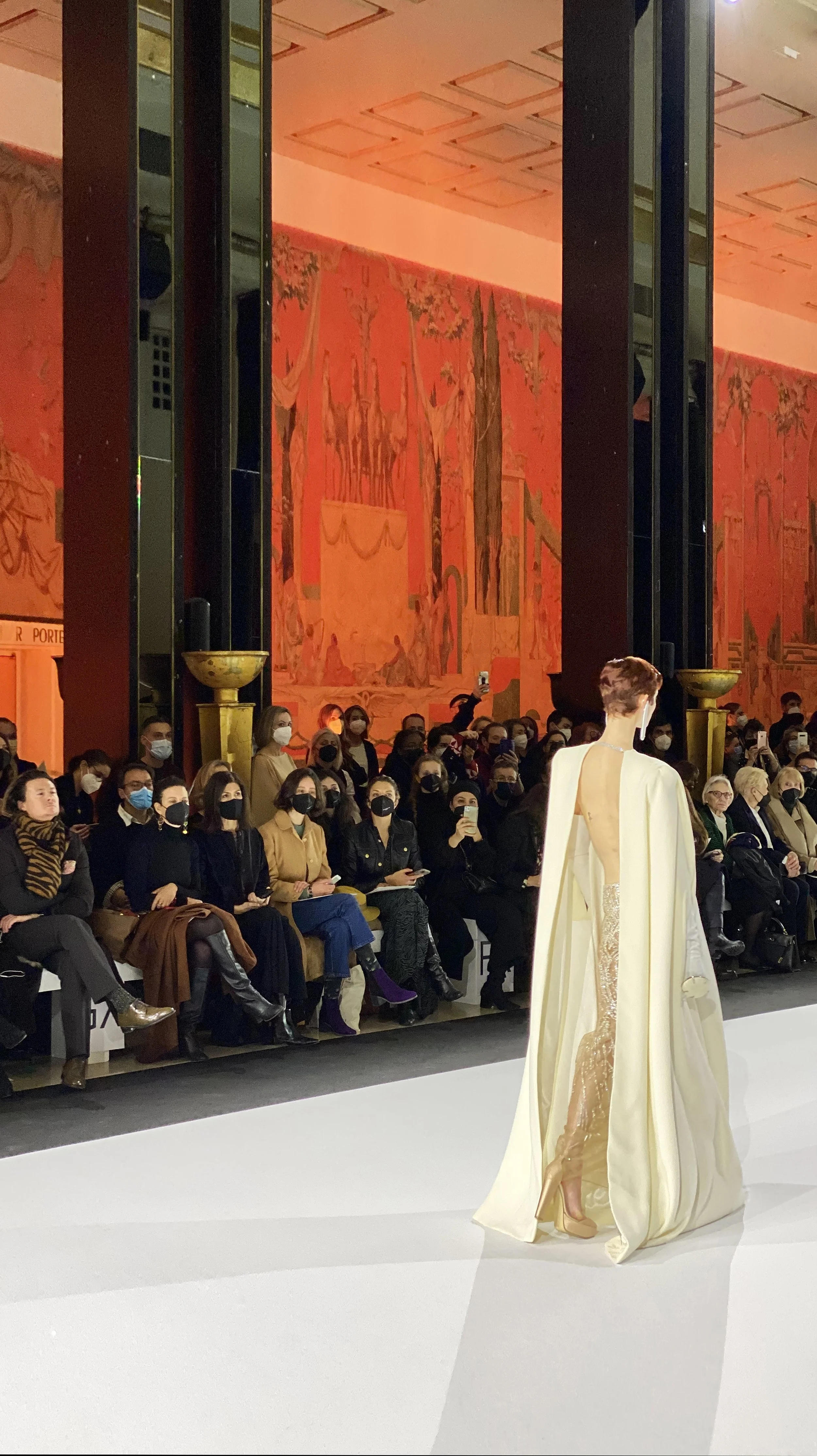 Fashion model walking on runway at a fashion show audience seated on either side, many wearing face masks, large wall art with red and orange hues in background.