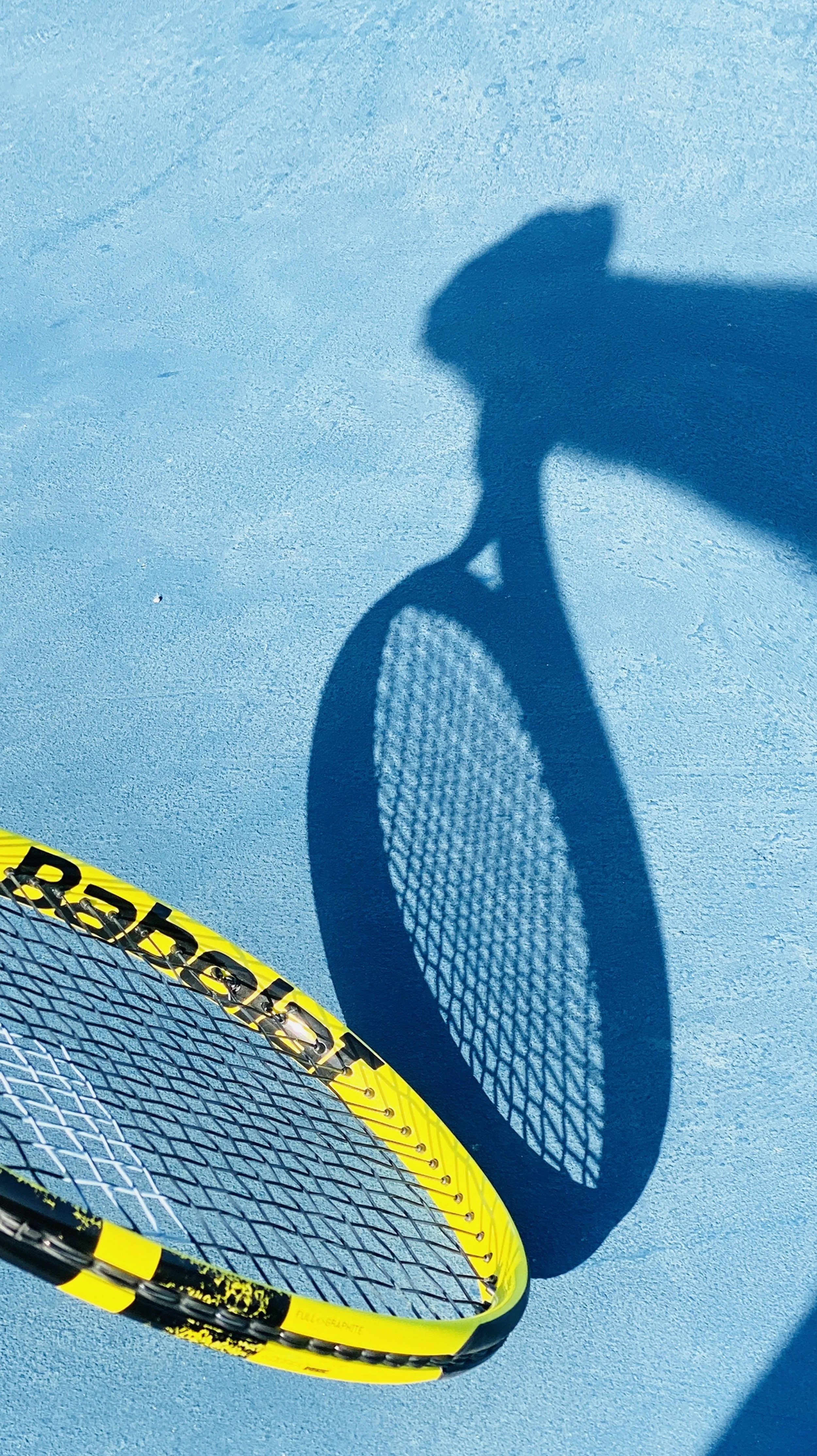 Shadow of a tennis racket on a blue court, with part of the racket visible in the bottom left corner.