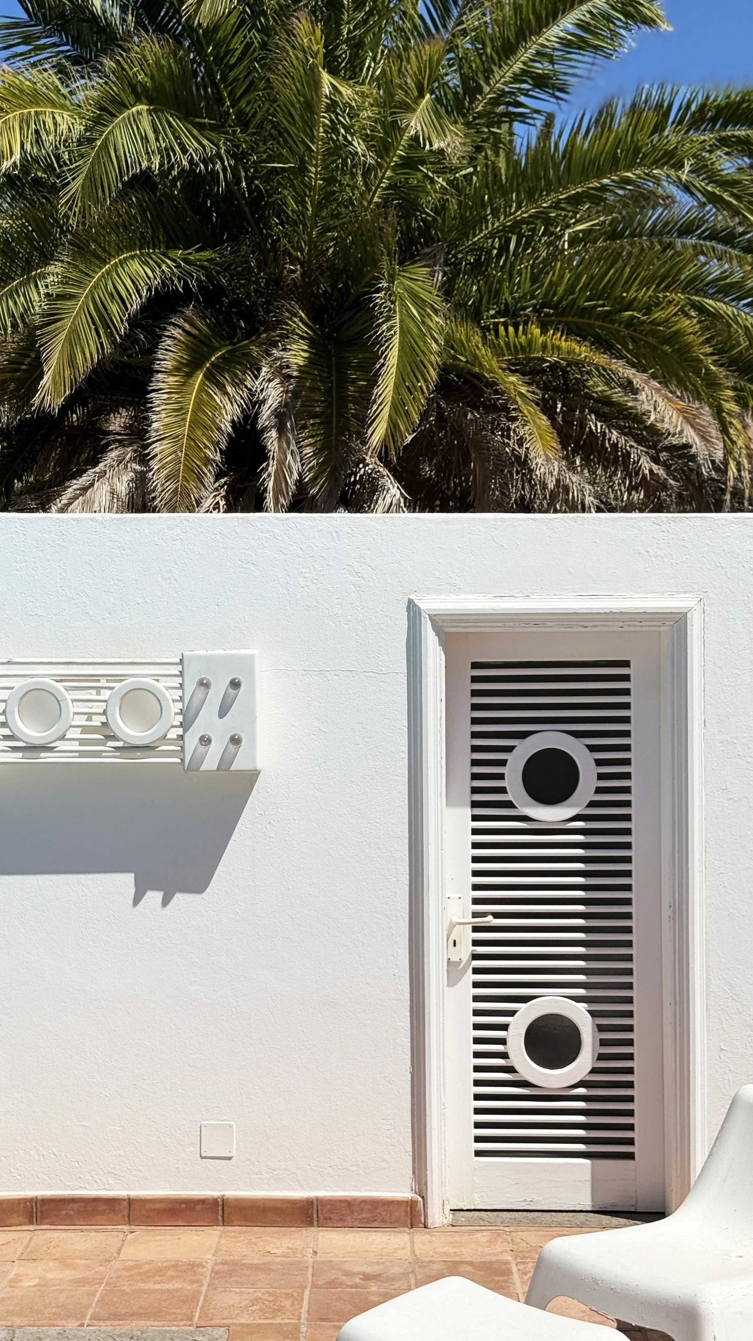 A white exterior wall with a door featuring horizontal slats and two round vents, a white wall-mounted heater or vent, terracotta tiled floor, partially visible white chair, and a large palm tree with green fronds against a blue sky in the background