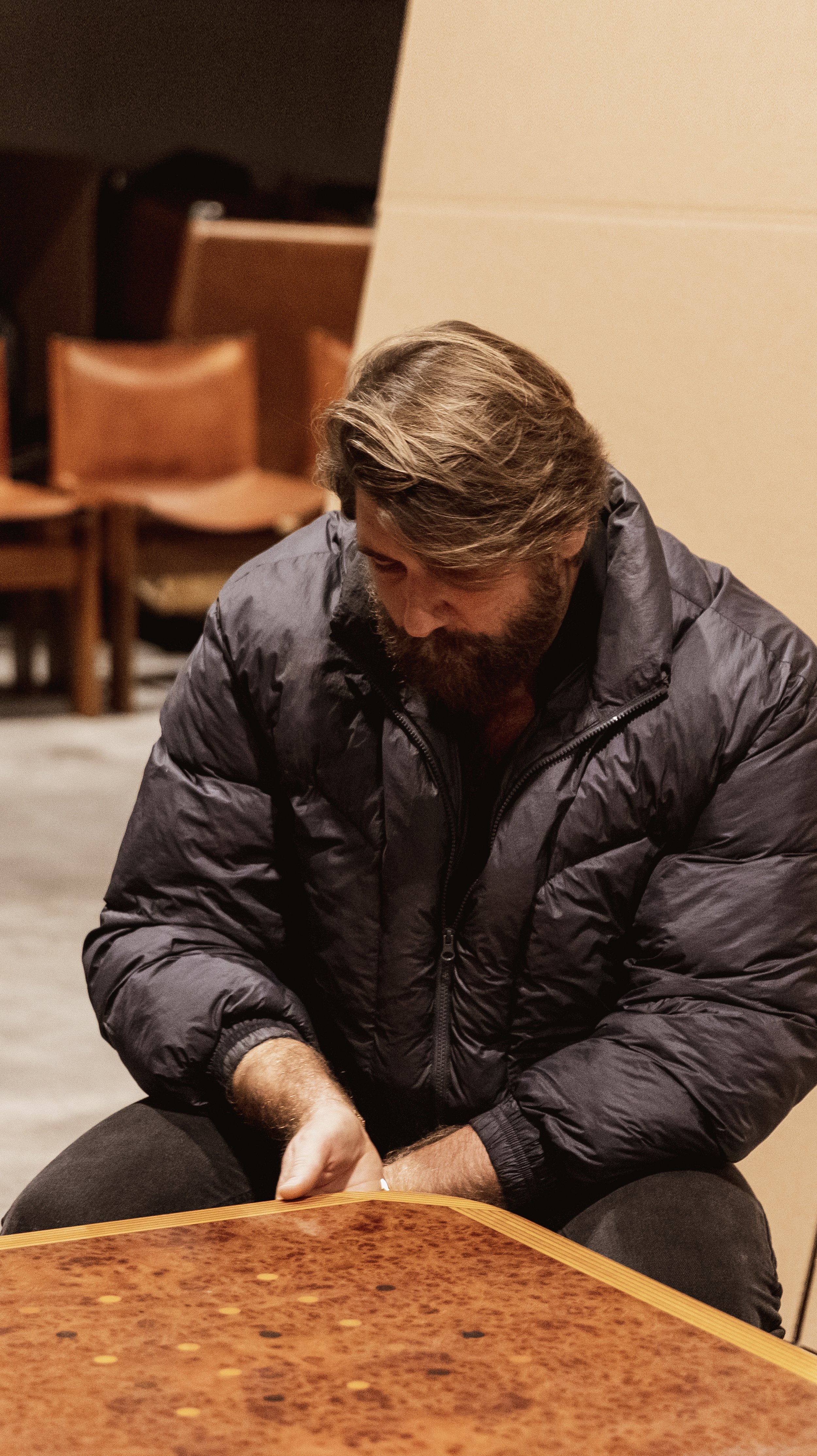 A man with a beard and long hair wearing a black jacket sitting at a table.