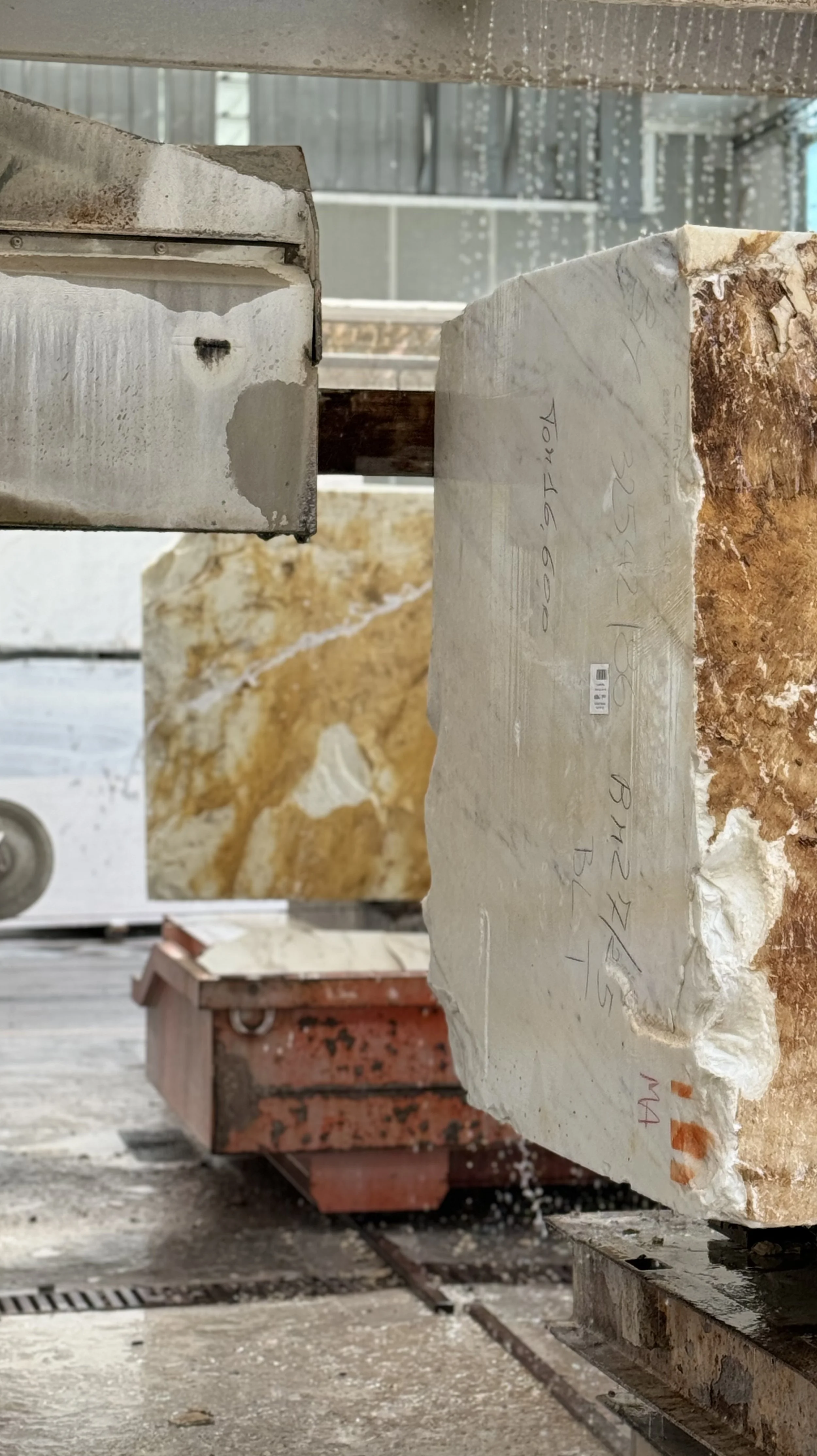 A close-up of a large, rough-edged slab of light-colored stone or marble, with handwritten markings and labels, in a workshop or warehouse setting.