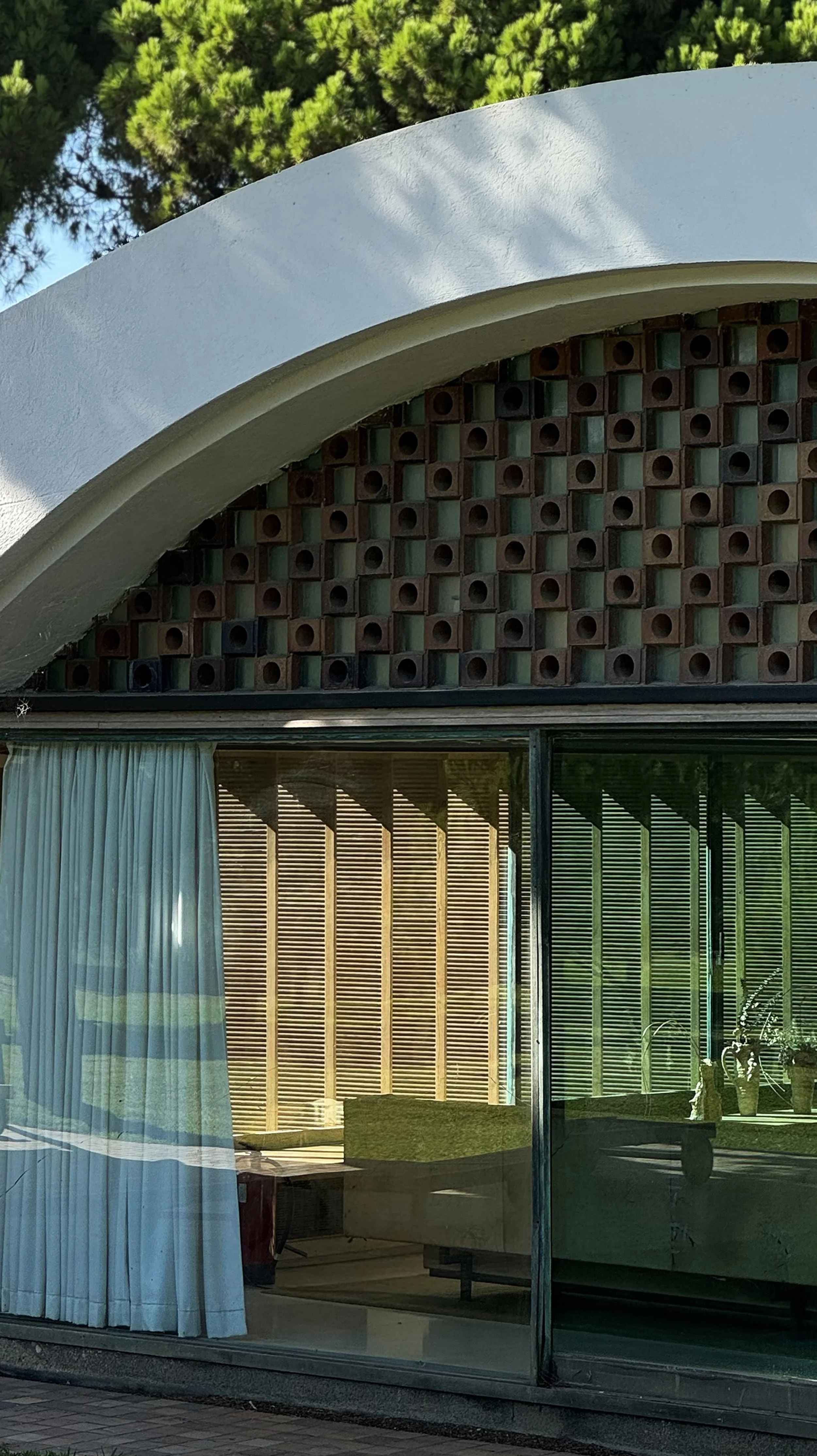 Close-up of a modern building with a curved white roof, a decorative brick wall with circular openings, and large glass windows reflecting outdoor surroundings.