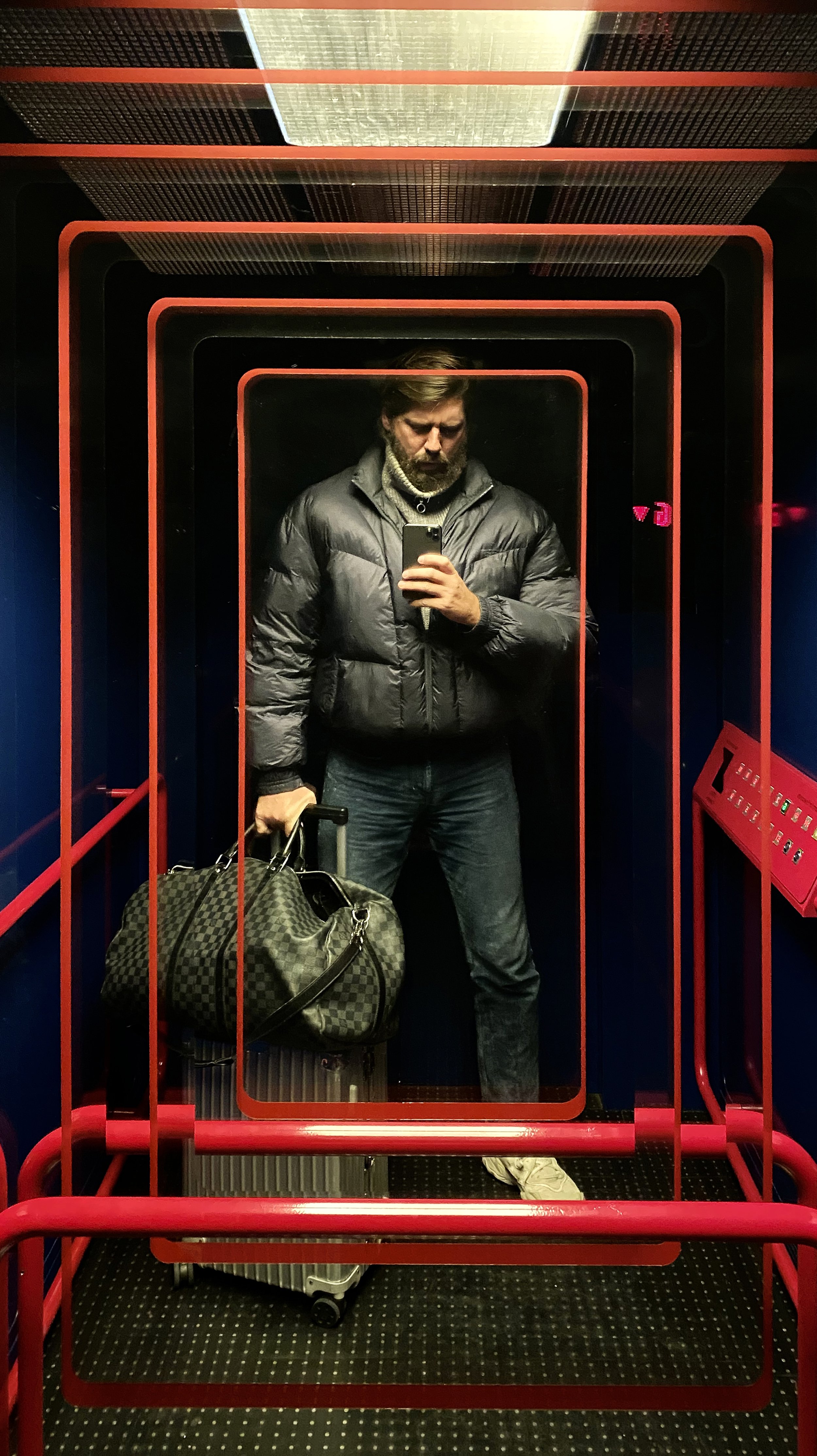 A man taking a selfie in an elevator mirror, holding a rolling suitcase and a checkered duffel bag, wearing a gray jacket, jeans, and white shoes.