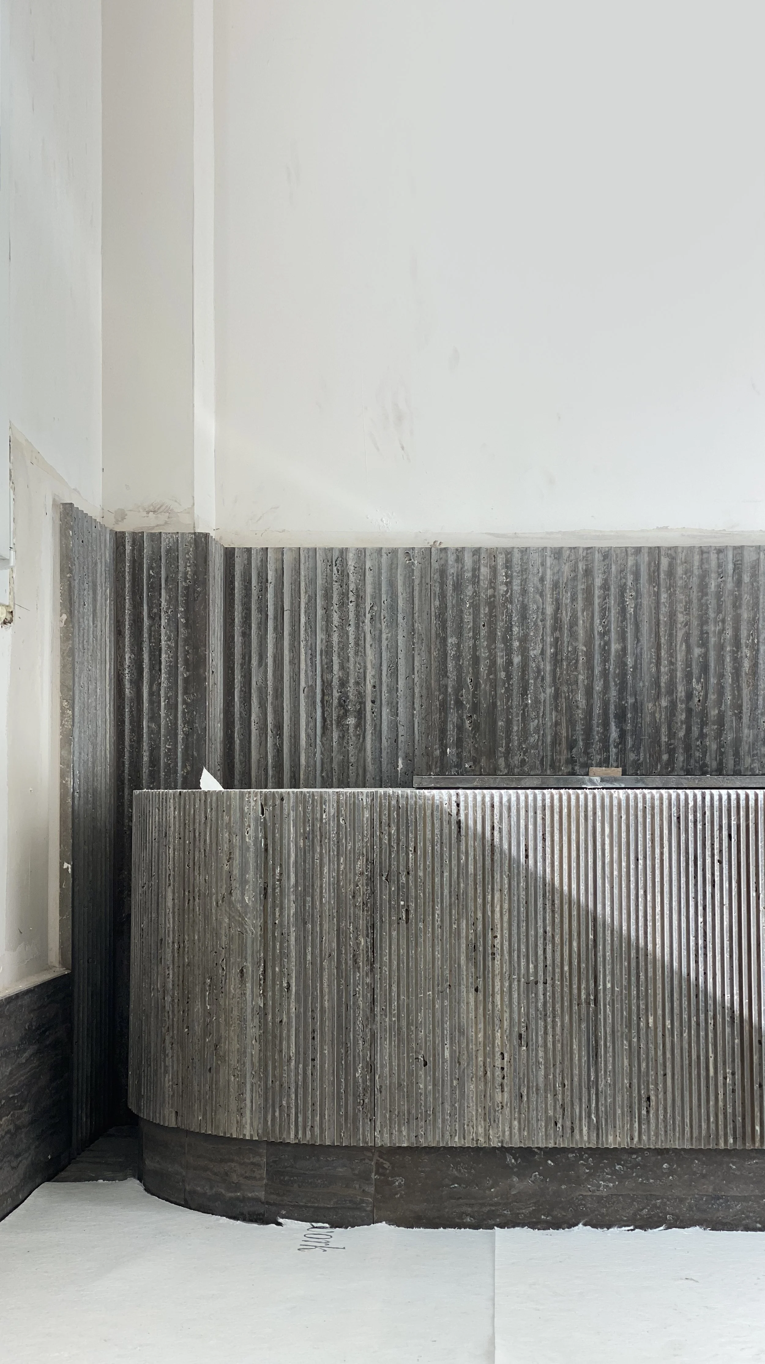 Close-up of a textured, corrugated metal surface forming a curved reception desk or counter, with a plain white wall in the background.