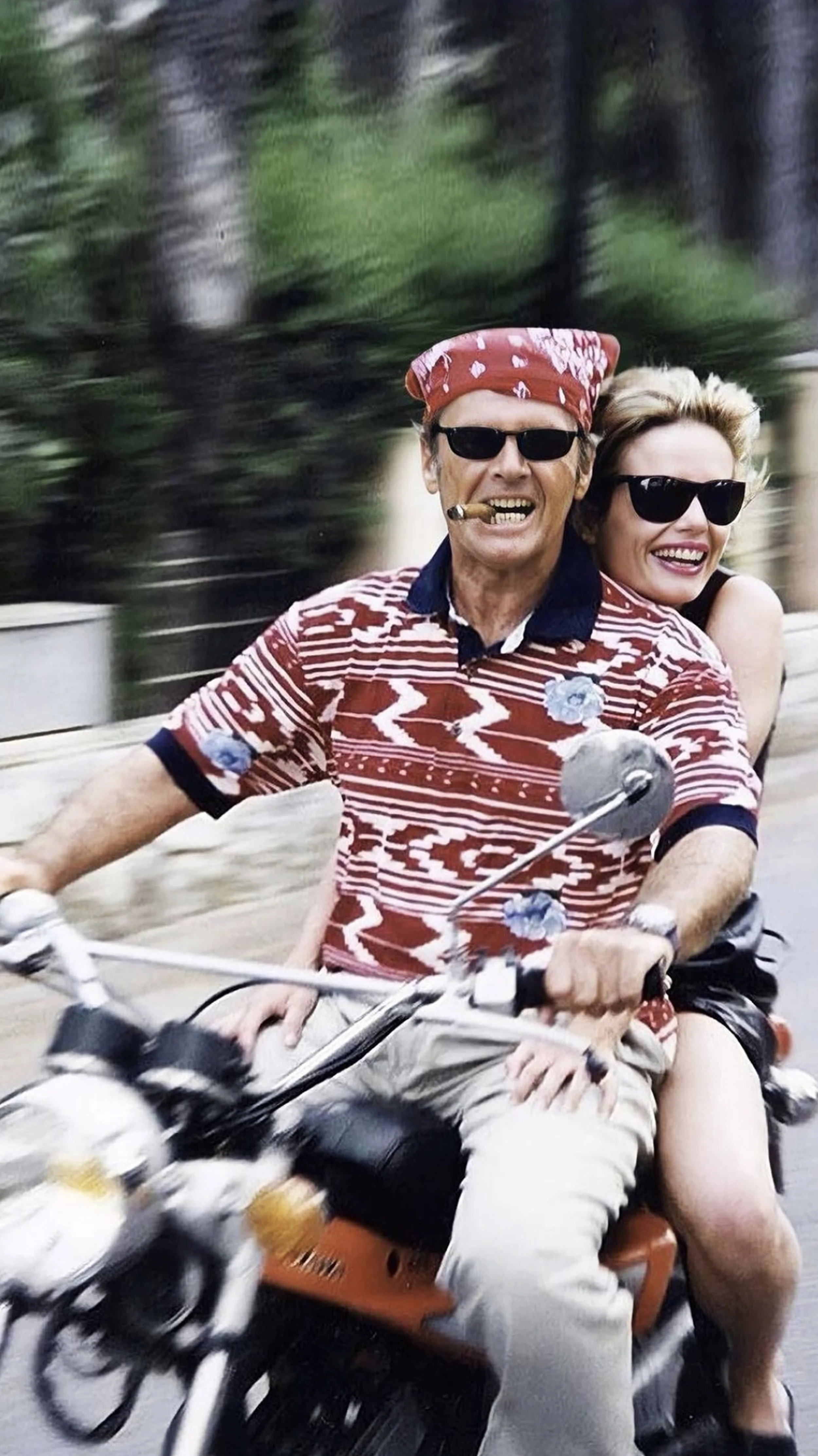 Two people, a man and a woman, riding a motorcycle together. The man is wearing sunglasses, a red bandana, and a patterned red and white shirt, smoking a cigar. The woman behind him is smiling, wearing sunglasses, and has blonde hair. They appear to 