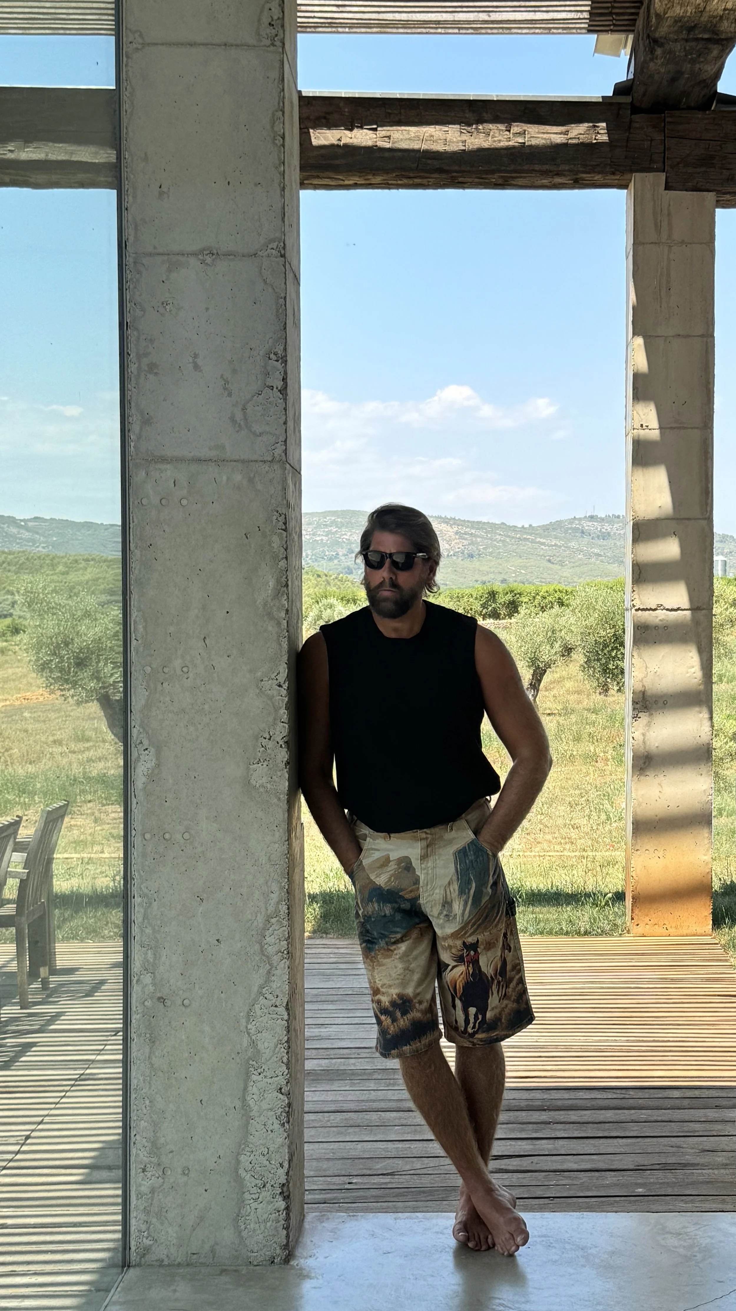 A man with dark hair and beard wearing sunglasses, a black sleeveless shirt, and camel-colored shorts with a horse print, standing barefoot against a concrete wall and glass window with an outdoor landscape of green fields, trees, and hills in the ba