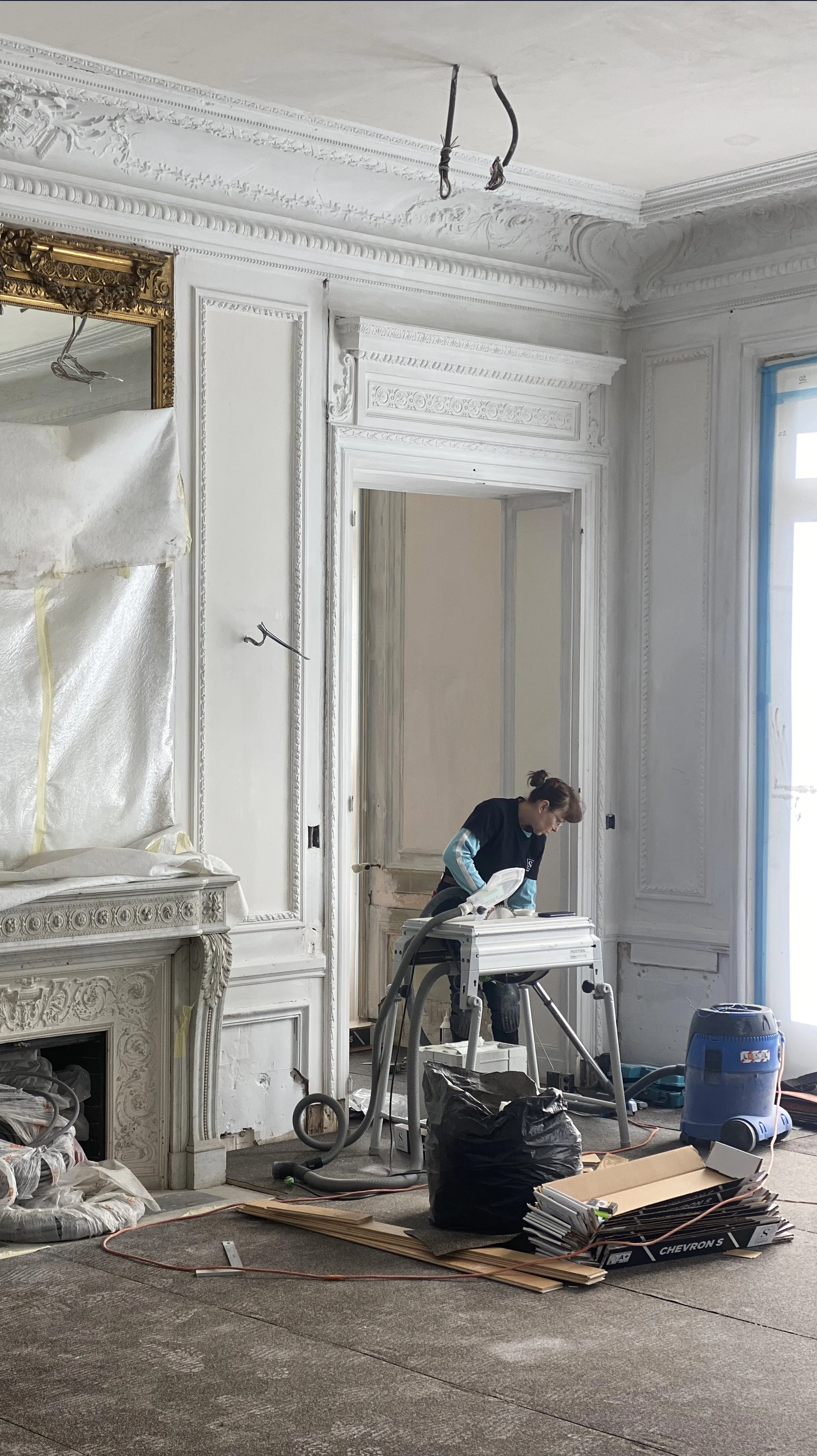 A worker with a power sander in a room with ornate white walls and a large window, which is covered in protective blue film. There are construction materials and equipment on the floor, including wood planks, a black garbage bag, and a vacuum cleaner