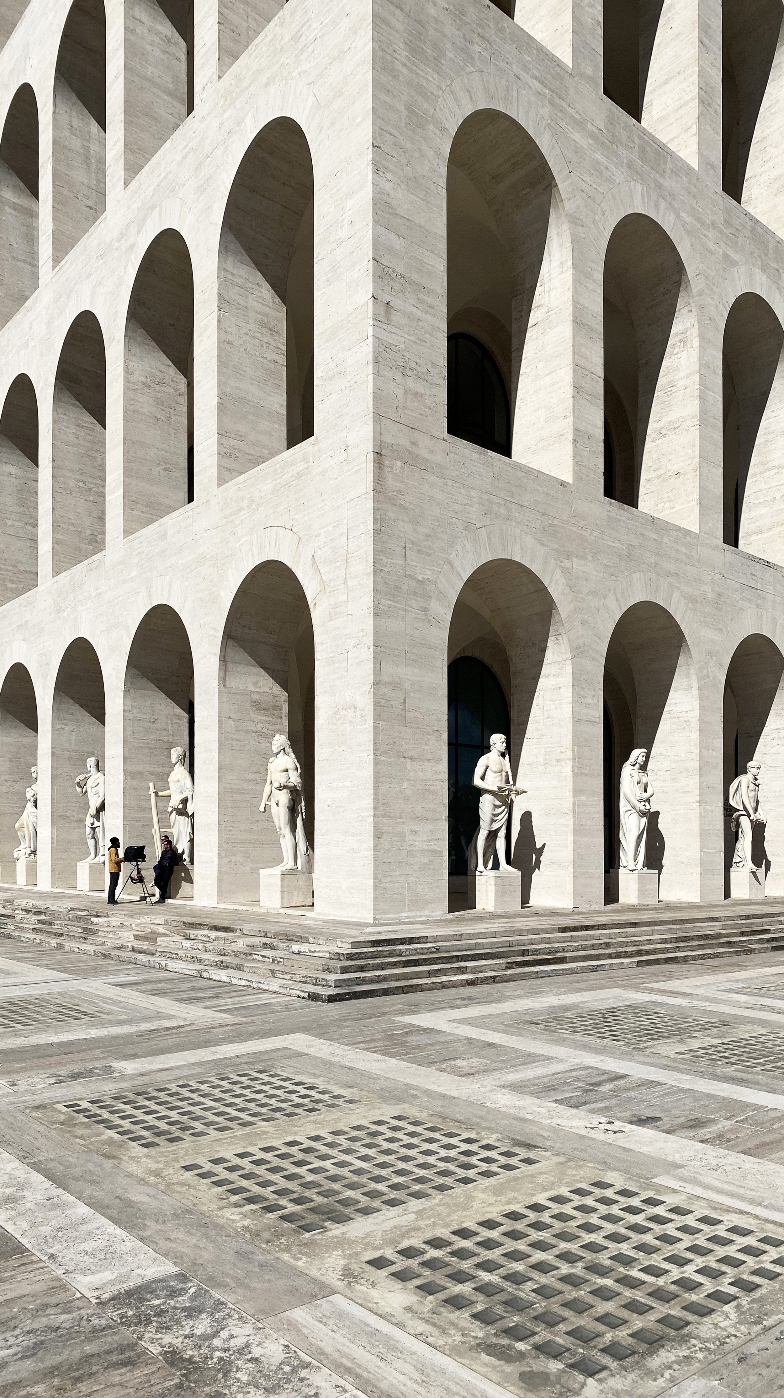 A large, classical-style building with multiple arched openings and statues along the front steps. Two people are near a camera set up on a tripod.