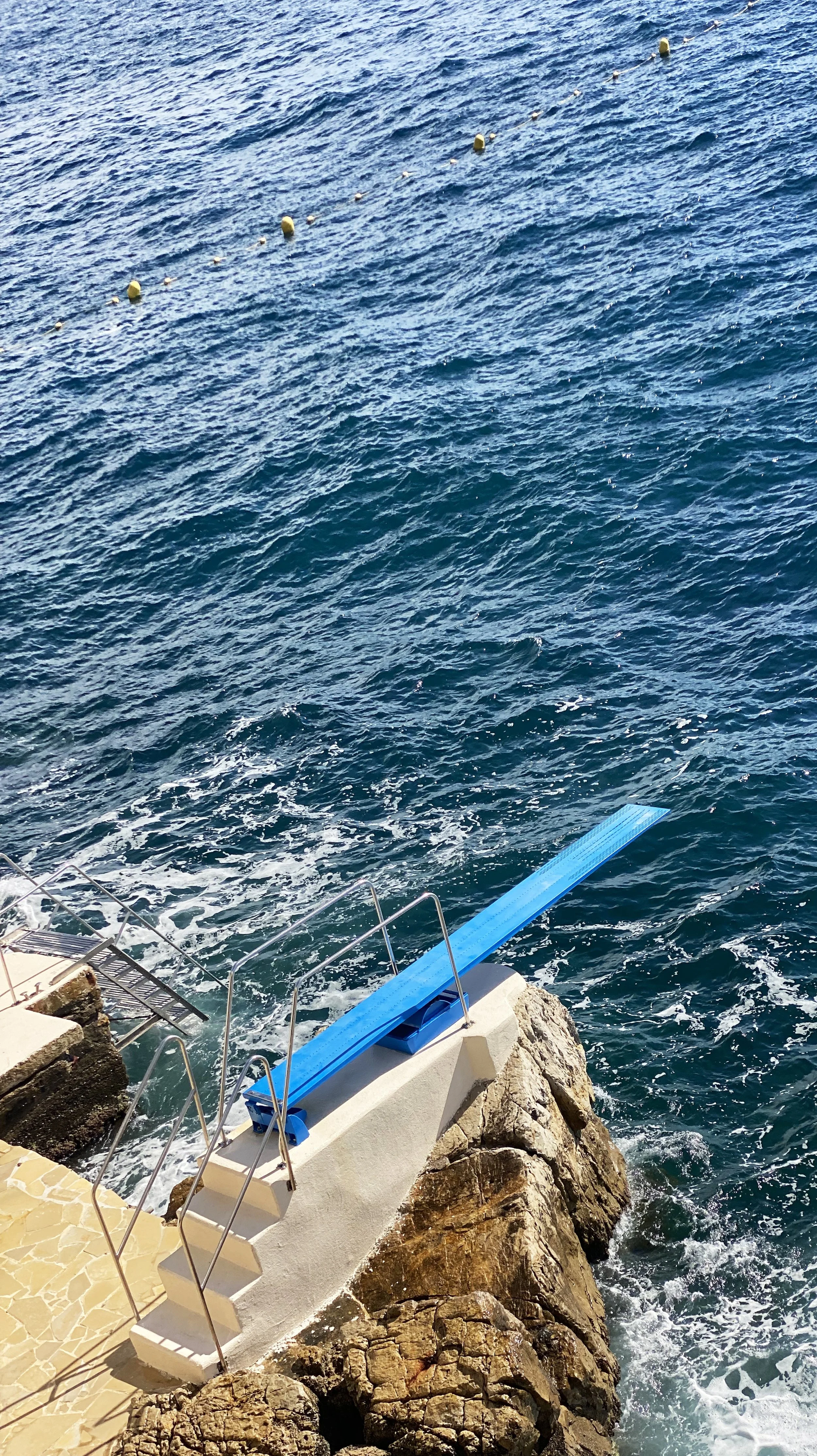 A blue diving board extends over rocky shore into the water near a seacoast with foam waves and distant yellow buoys.