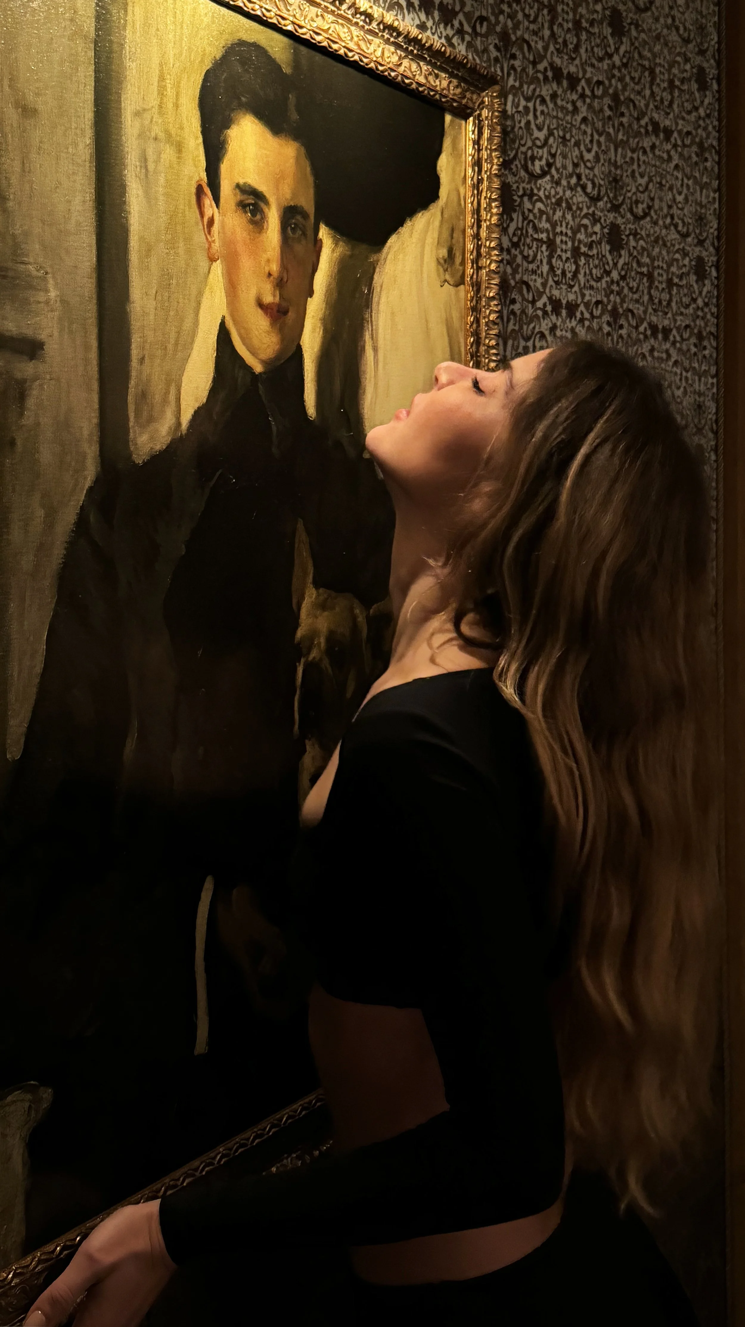 A woman with long wavy hair wearing a black dress standing near a framed oil portrait of a man in formal attire on a patterned wall.