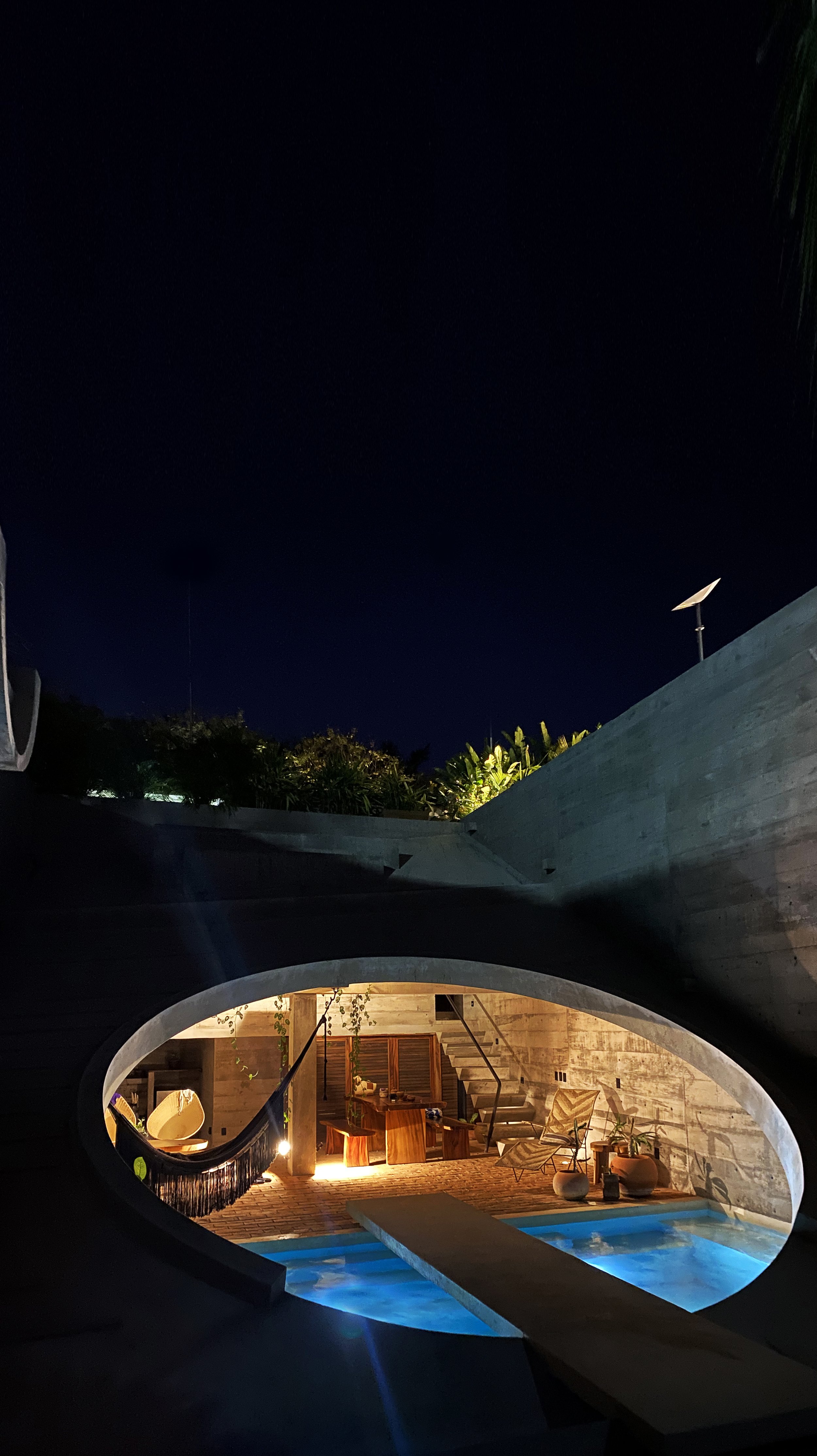 Nighttime view of a modern outdoor pool area with warm lighting, seating, and wooden furniture, framed by curved concrete structures and lush green plants.