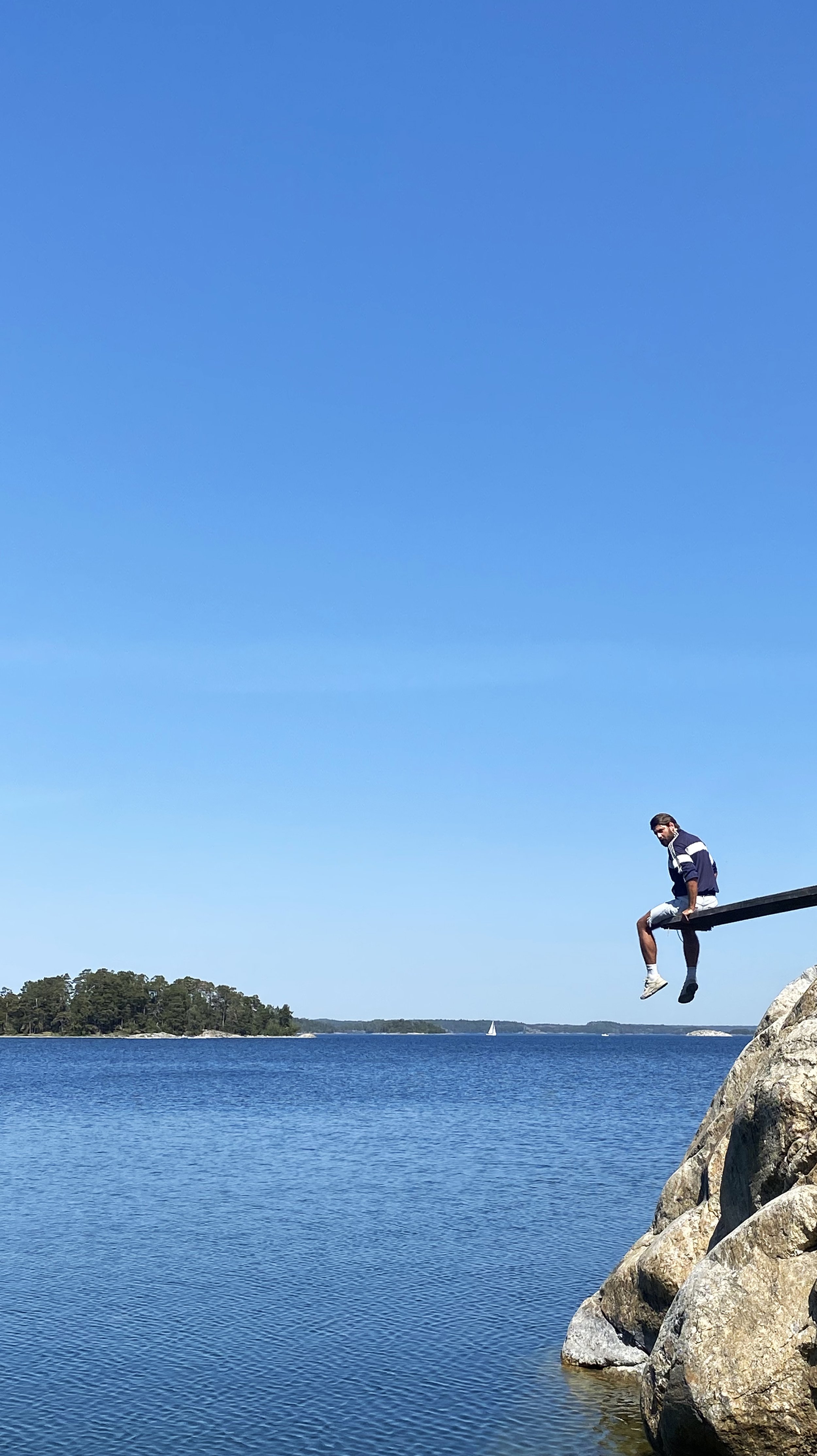 A young man sitting on a wooden beam extending over a rocky shoreline by a body of water on a clear, sunny day. In the background, there are trees on the opposite shore and a sailboat in the distance.