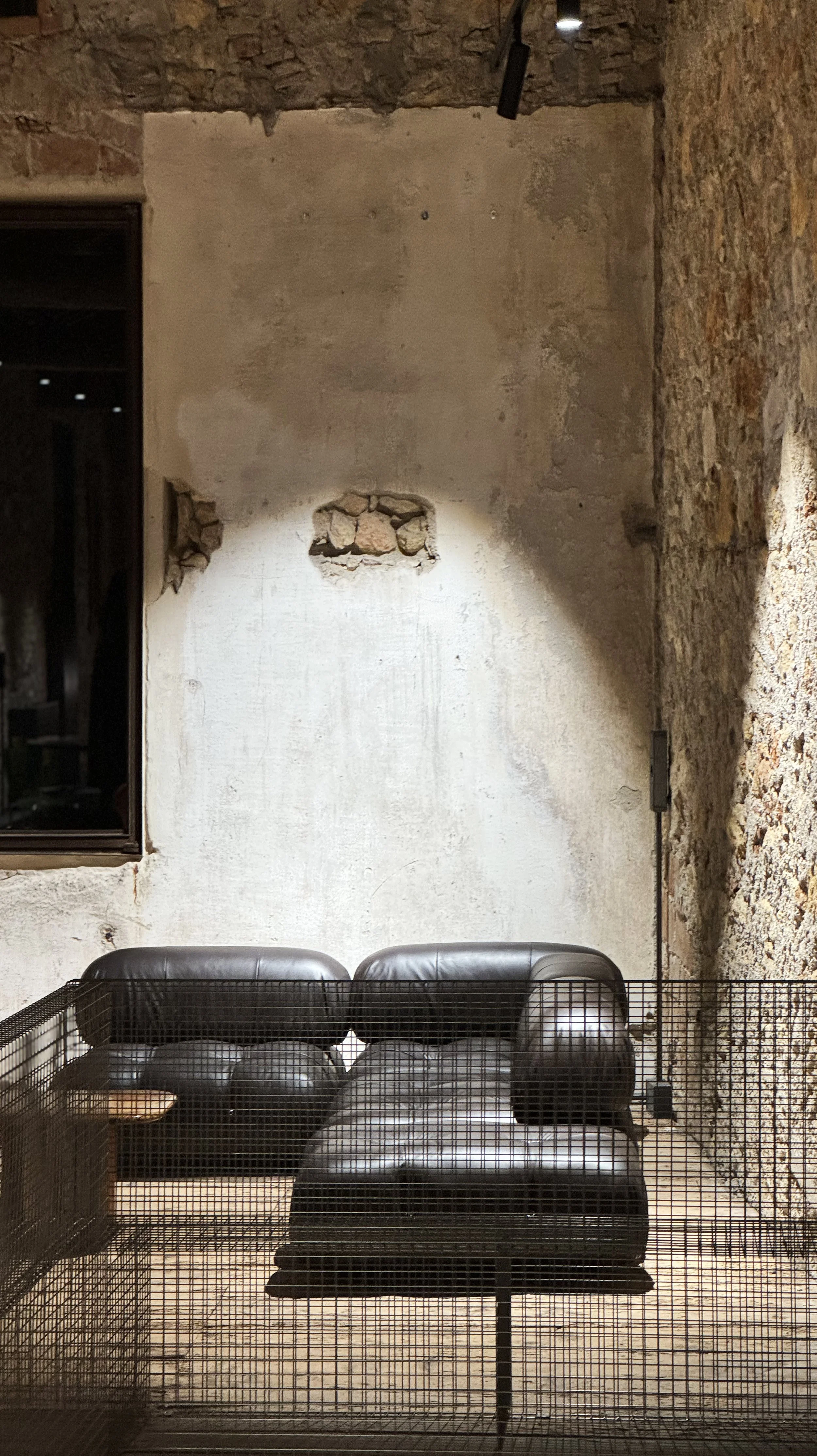 A black leather loveseat in front of a rustic, exposed brick and plaster wall.