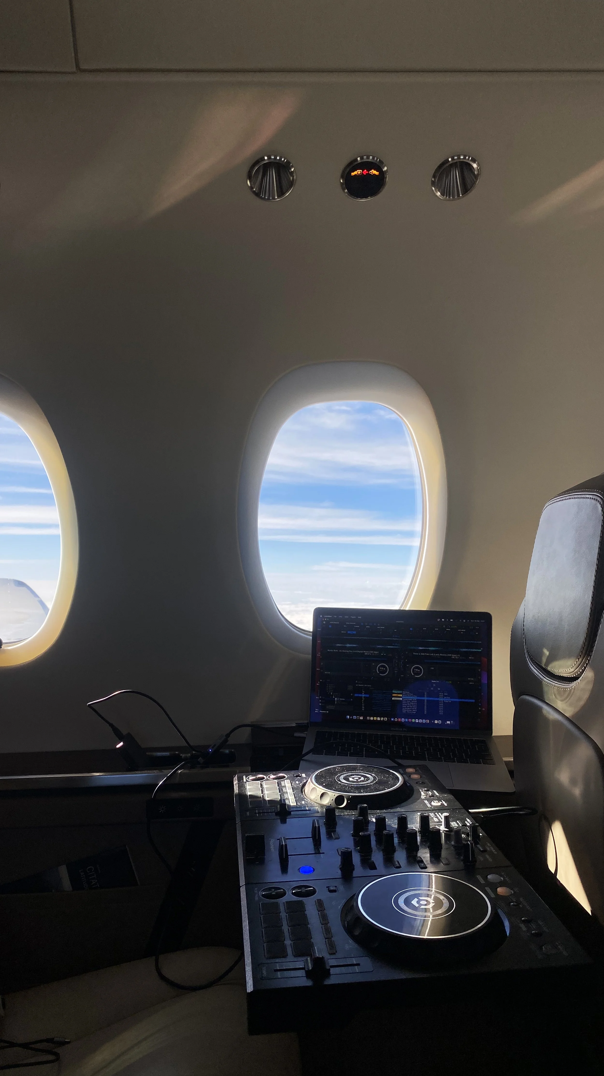 DJ equipment set up on a tray table in an airplane with a laptop and sky visible through airplane windows.