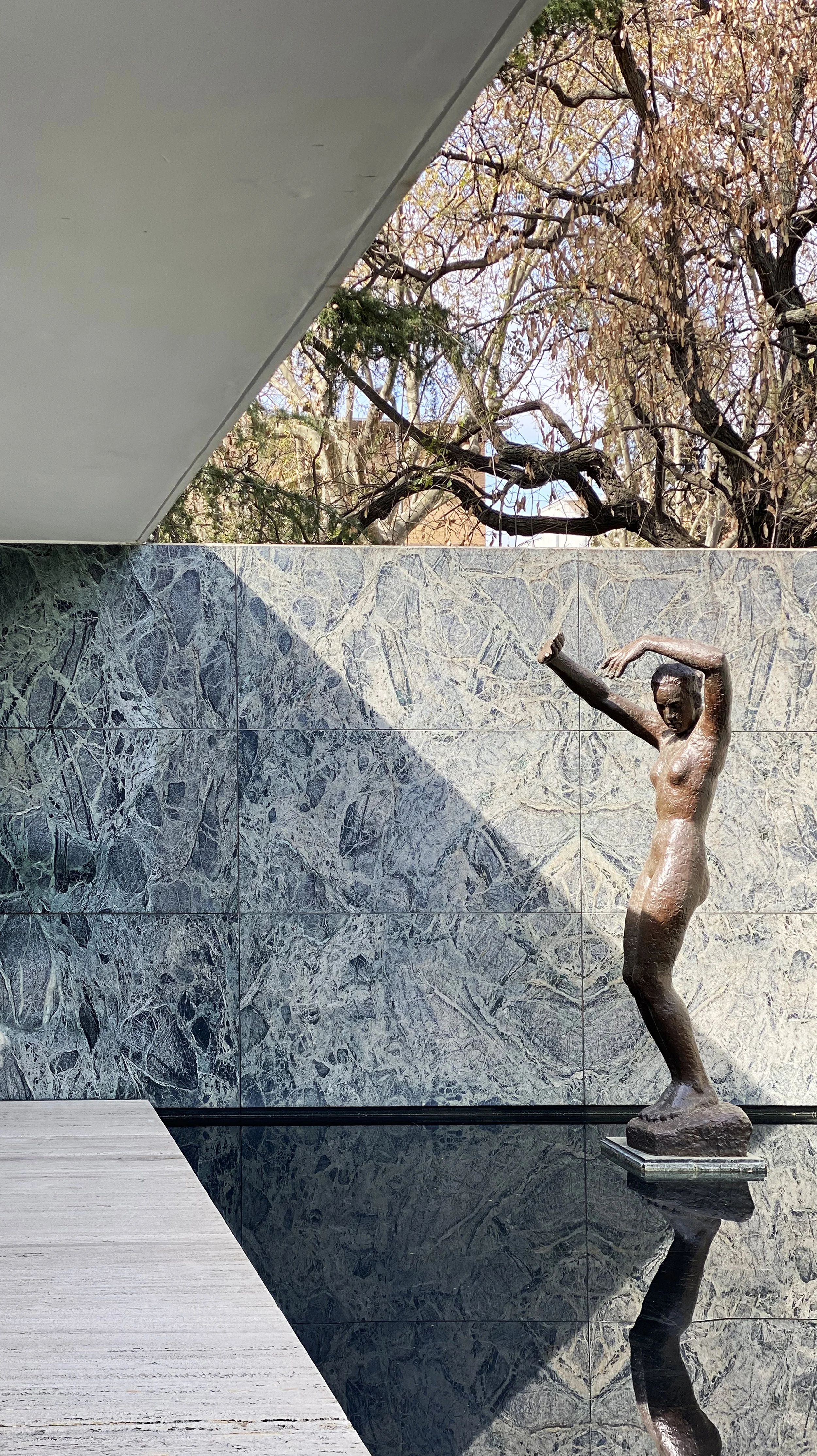 A bronze sculpture of a woman with arms raised above her head, standing on a rectangular base, reflected in a black reflective surface, with a decorative stone tile wall and autumn trees in the background.