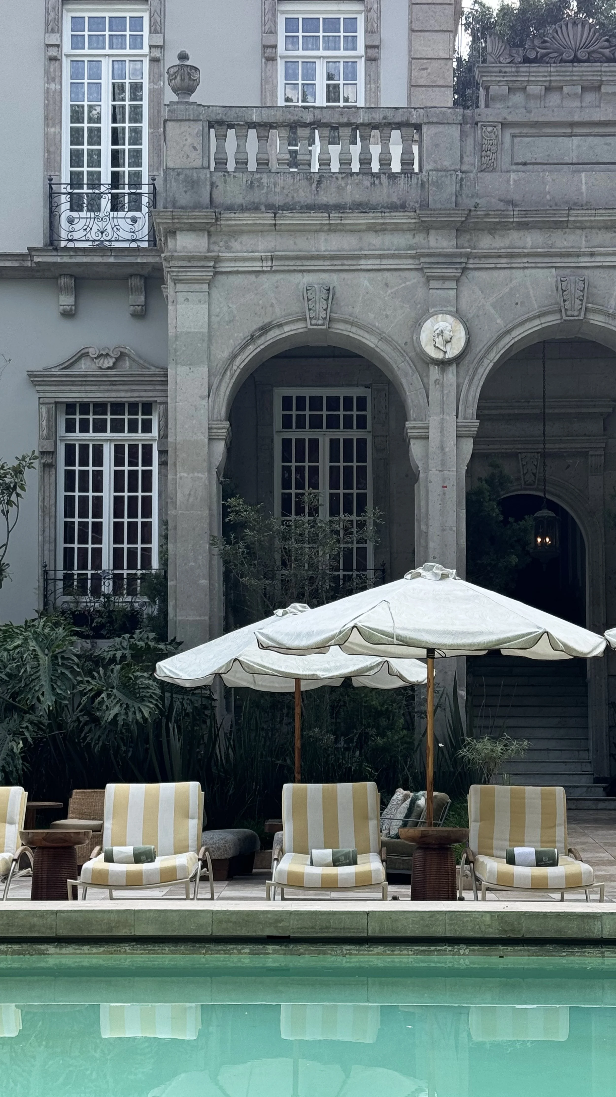 Chairs and umbrellas by a swimming pool in front of a grand stone building with large windows and arched entrance.