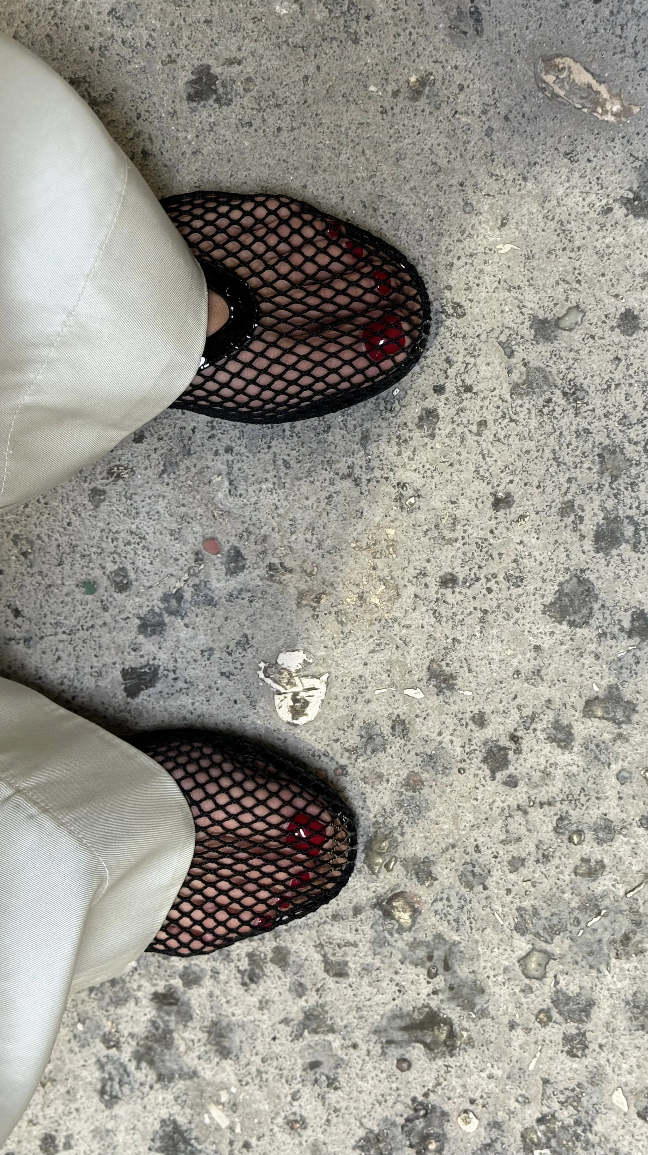 Person wearing fishnet shoes with red toes, khaki pants, standing on a rough concrete surface.